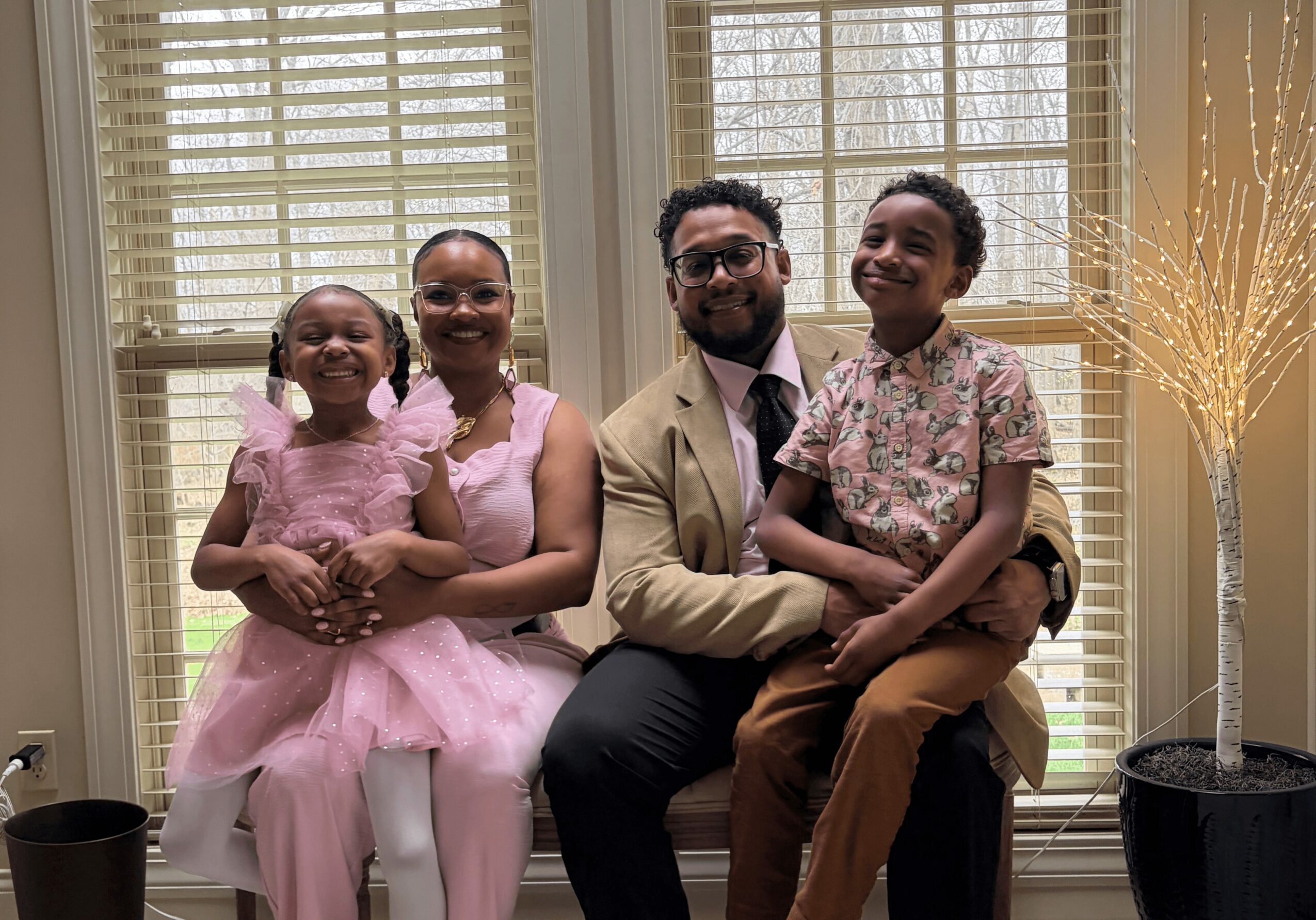 Four people sitting together indoors, smiling, with large windows and curtains behind them.