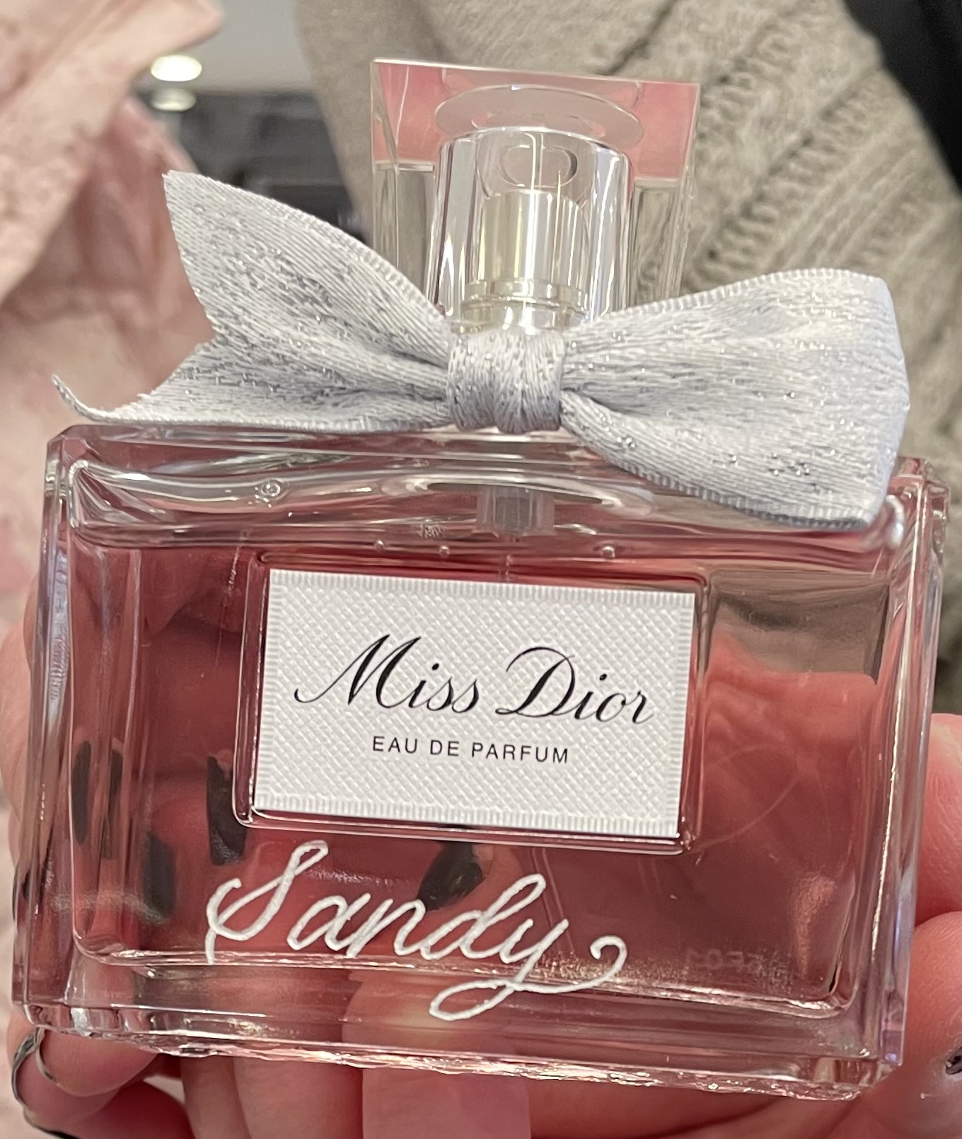 Clear perfume bottle with a white bow, pink liquid inside, label reads Miss Dior Eau de Parfum Sandy.