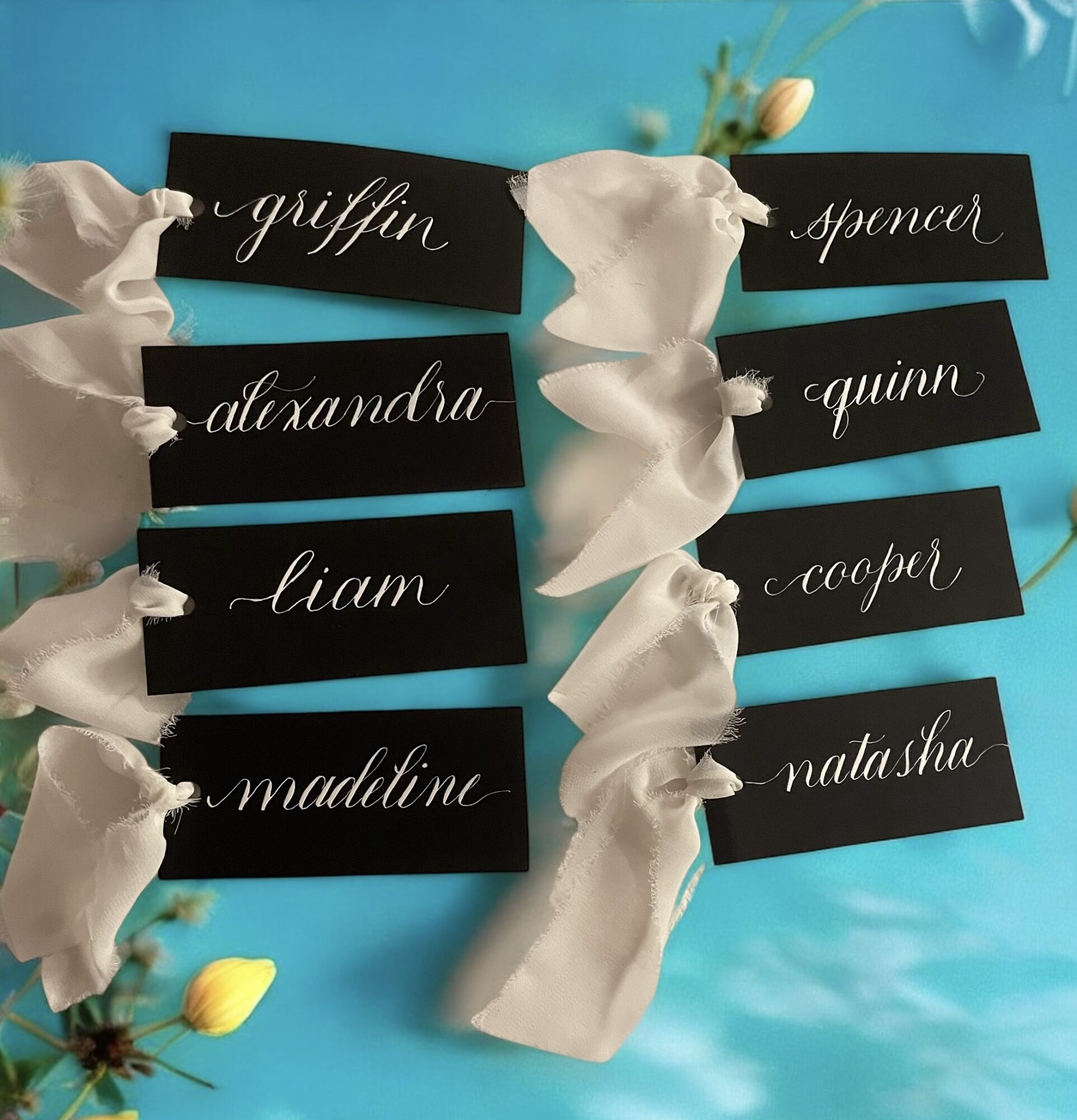 Black name tags with white cursive writing tied with white ribbons on blue background.