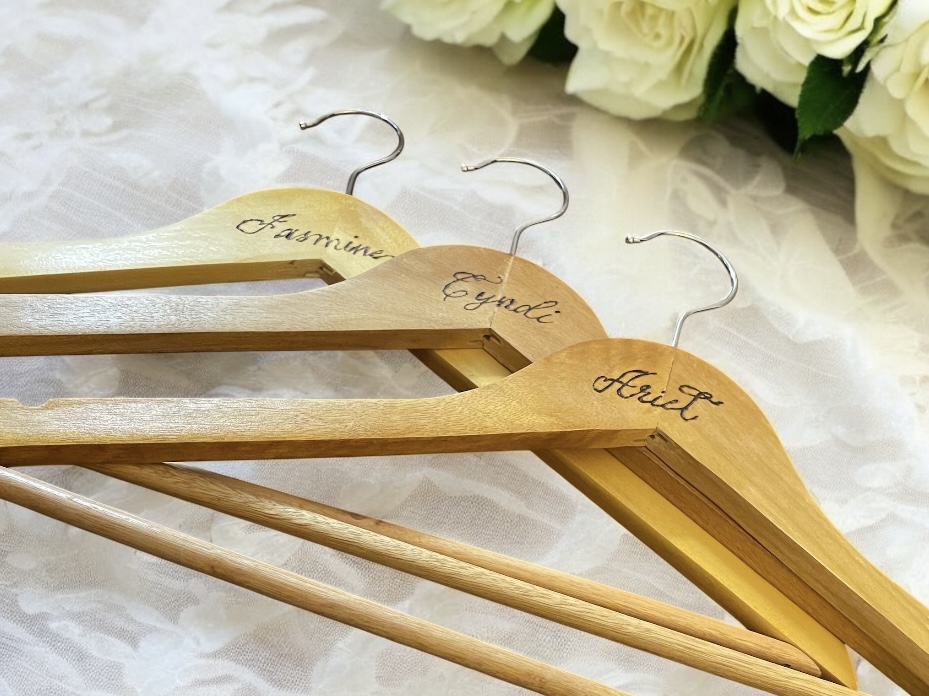Three wooden hangers with names written on them, placed on a white surface with white flowers in background.