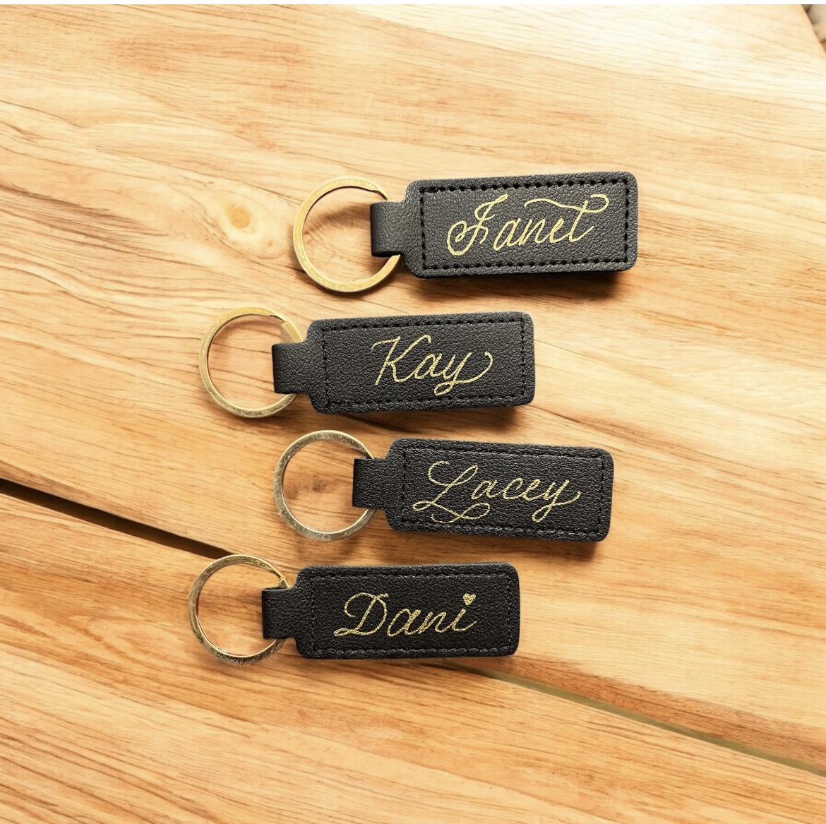 Four black keychains with gold cursive names: Fanet, Kay, Lacey, Dani, on wooden surface.