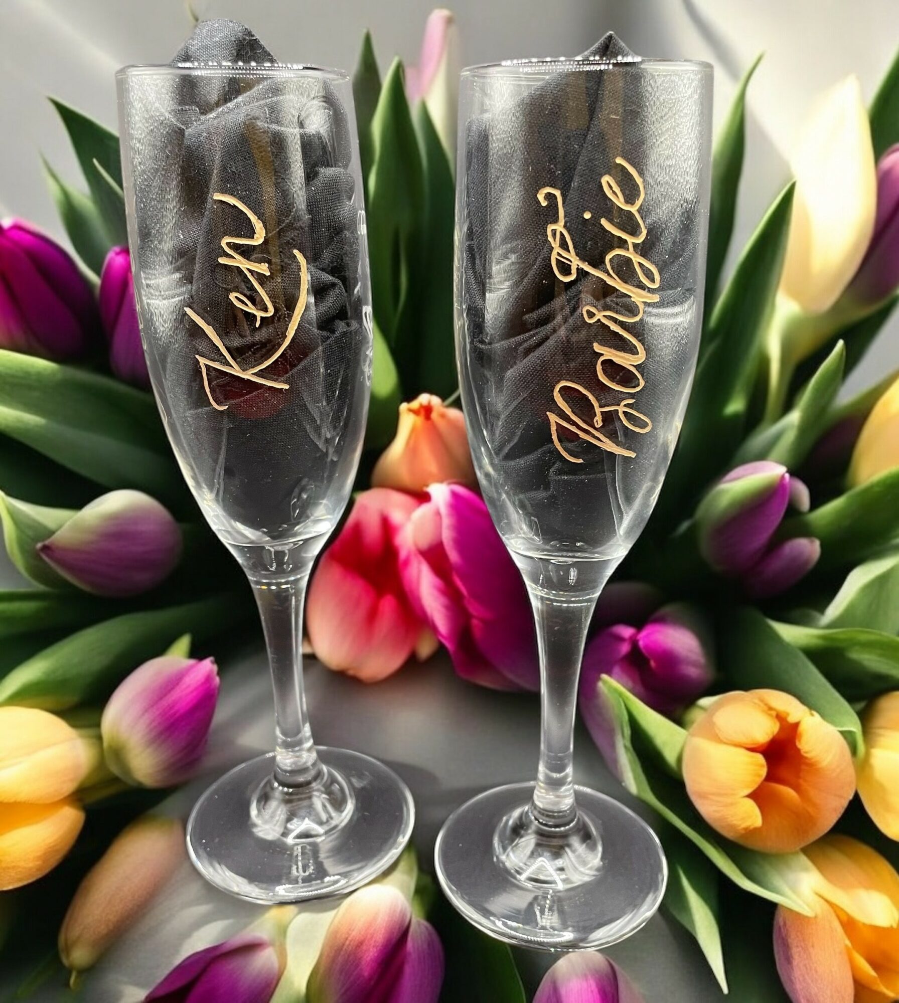 Two champagne glasses with names written on them, surrounded by colorful tulips and roses.