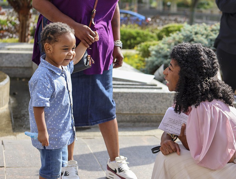 Young girl with braided hair smiling at woman kneeling and holding a paper outdoors.