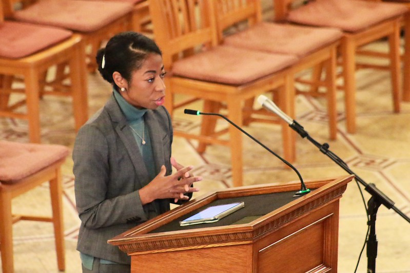 Woman speaking at a wooden podium with a tablet, surrounded by empty chairs in a room with wooden flooring.