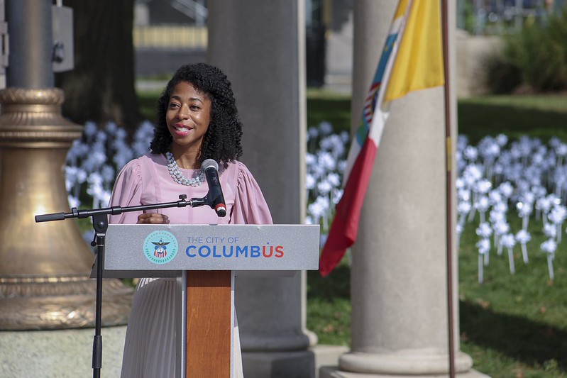 Woman speaking at podium outdoors with a flag and flowers in background.