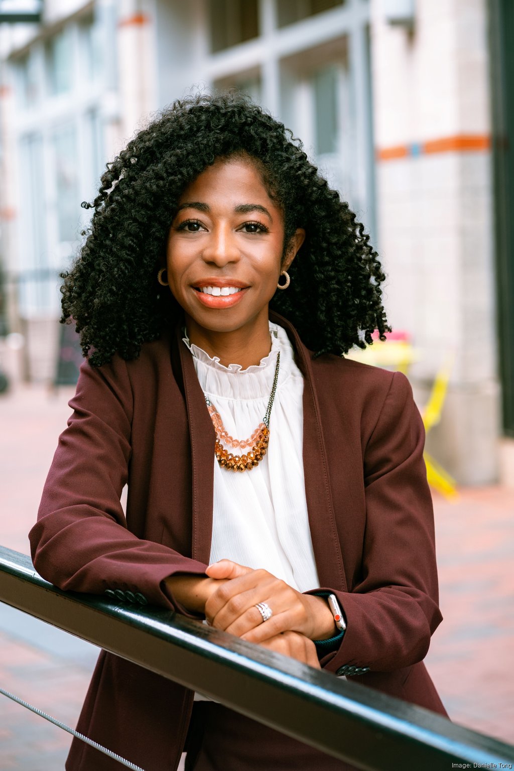 Woman with curly hair smiling, wearing a brown blazer and white blouse, outdoors in an urban setting.