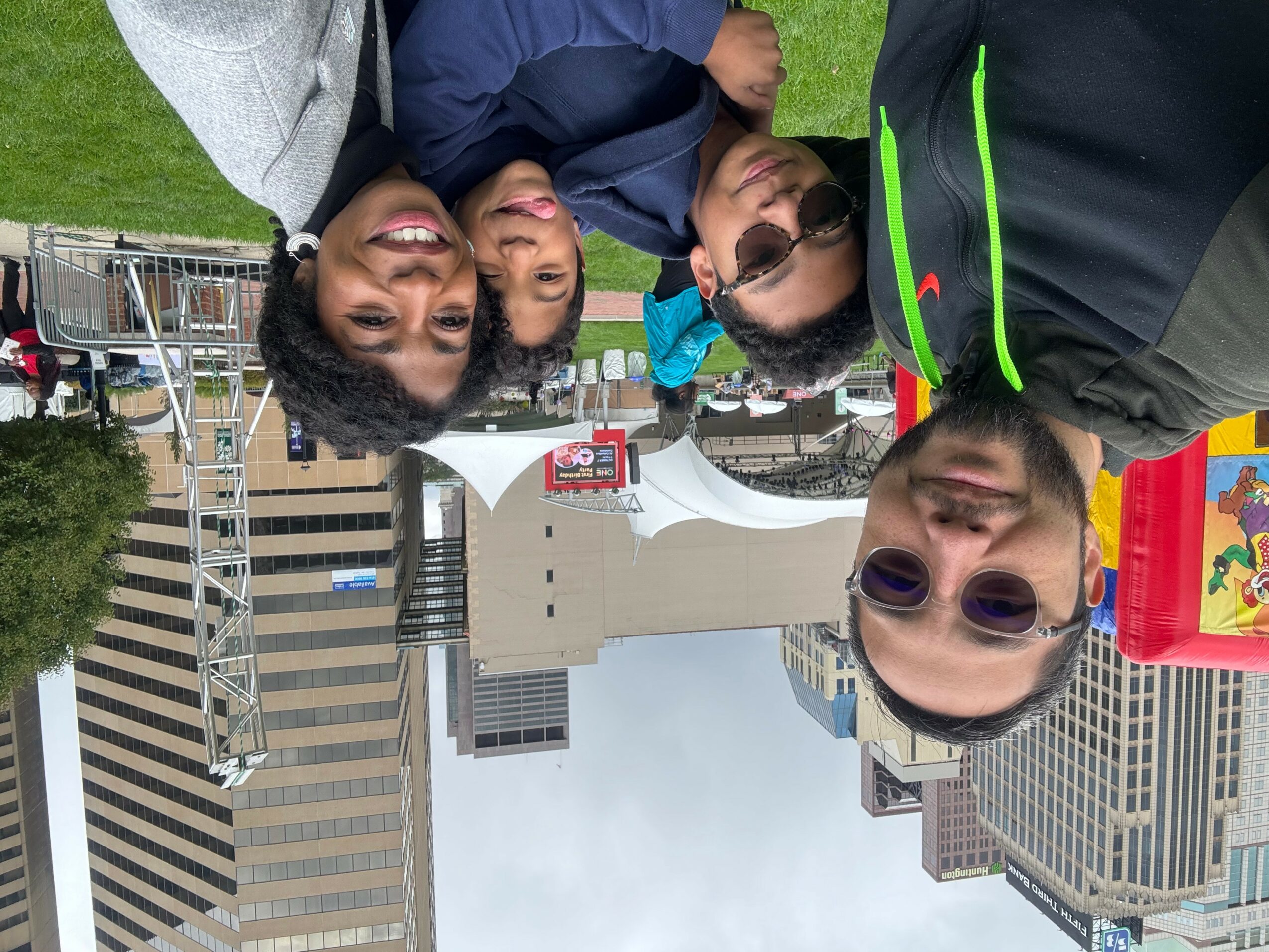 Group of four people smiling and posing outdoors in front of city buildings and a white structure.