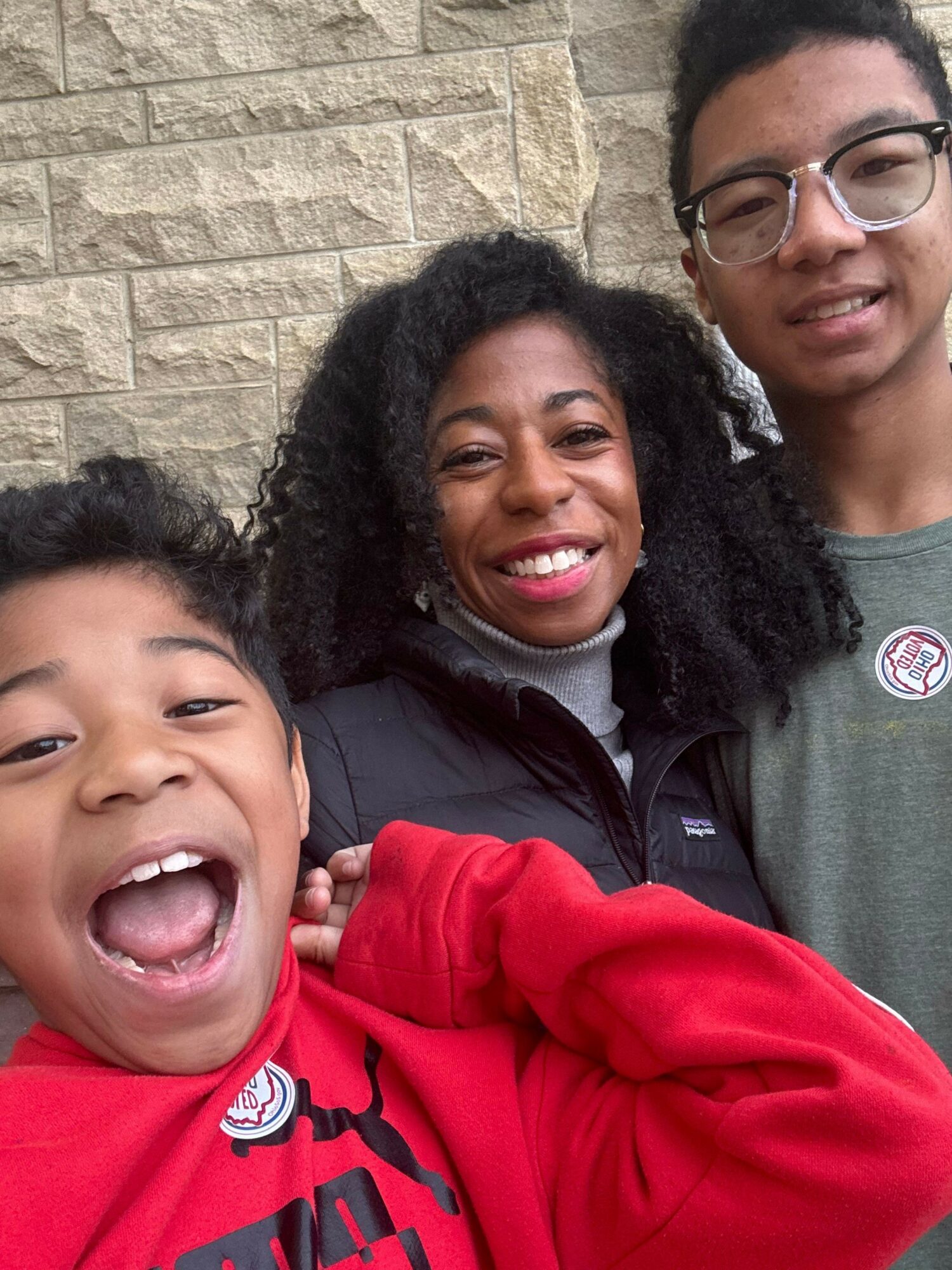Three smiling people close together, two women and one man, in front of a brick wall.