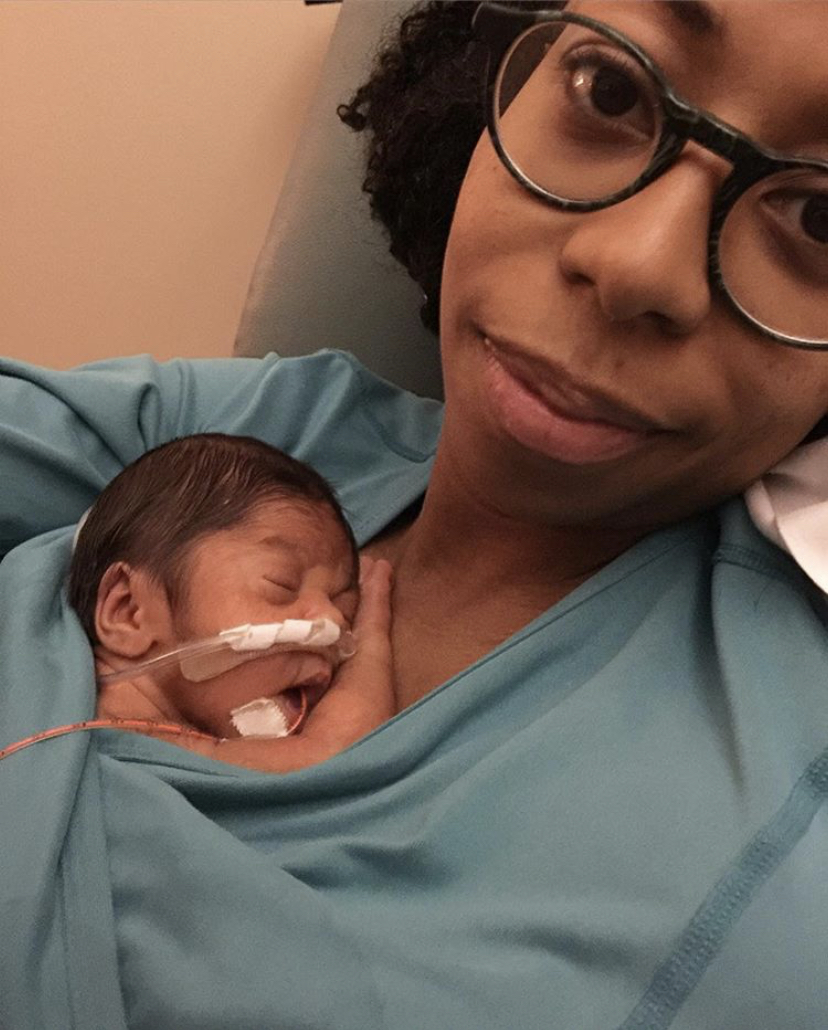 Person holding a newborn baby with a nasal tube, both wearing blue clothing, in a hospital setting.