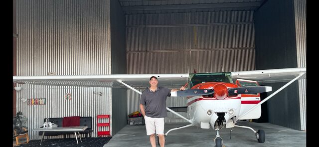 Person standing next to small aircraft inside hangar with tools and equipment nearby.