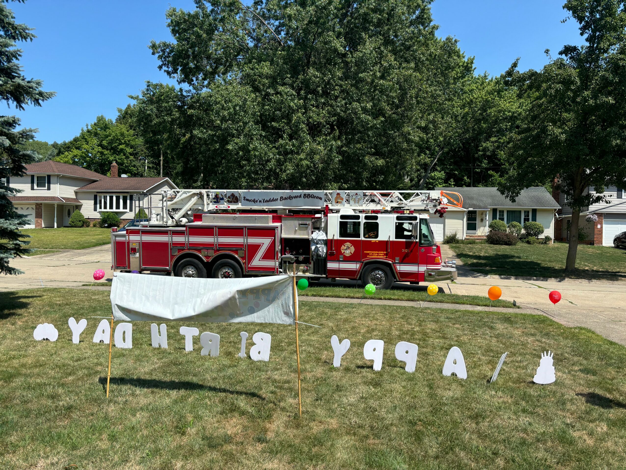 Fire truck parked on a lawn with trees and houses in the background, surrounded by colorful balloons and decorations.