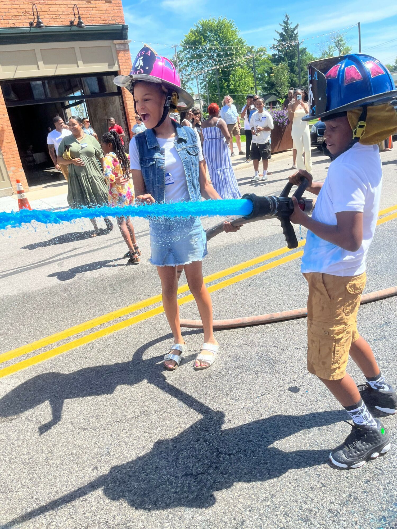 Two children wearing helmets and casual clothes play with a water hose on a street during a sunny day, with people in the background.