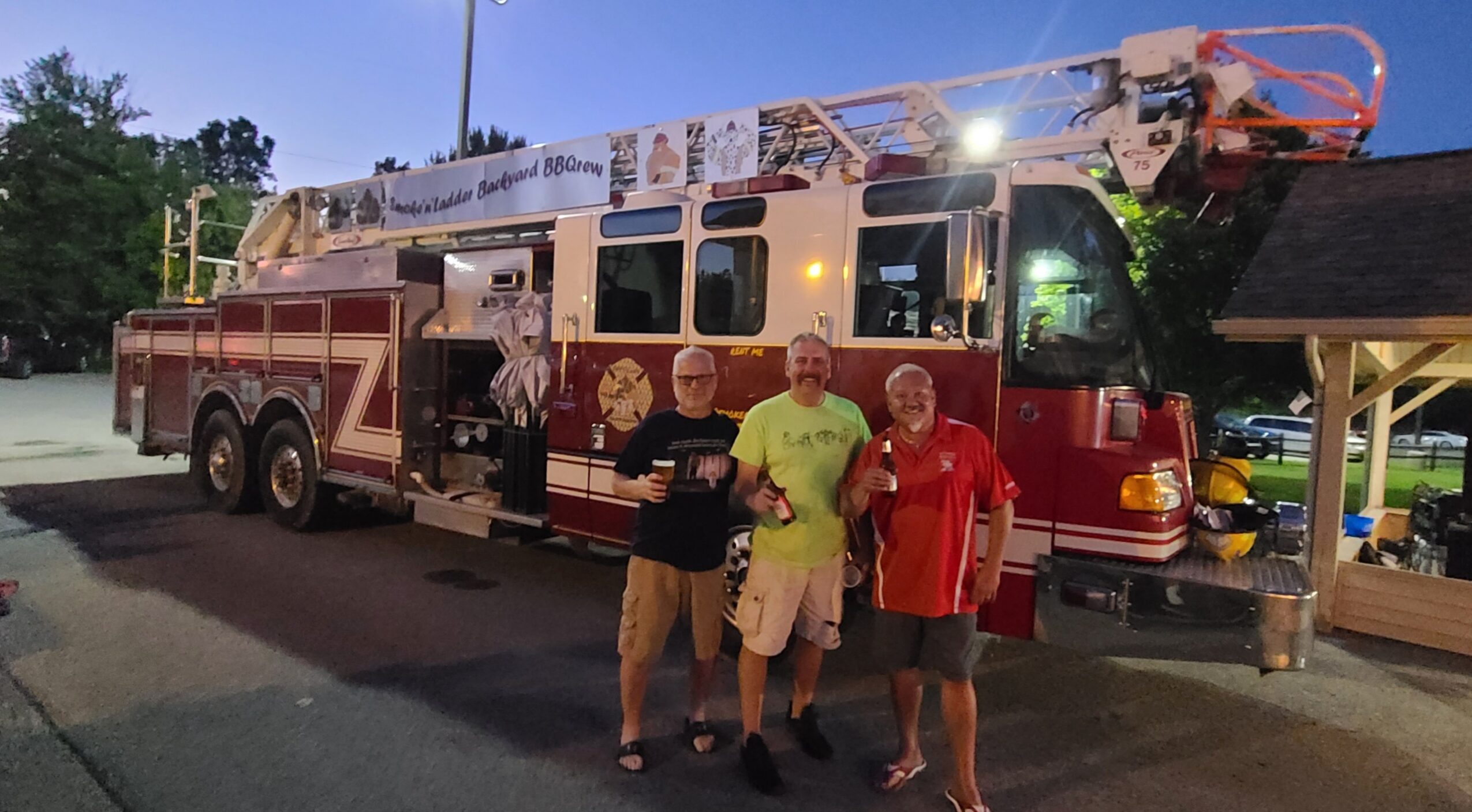 Three men stand in front of a fire truck at night, with trees and a building in the background.