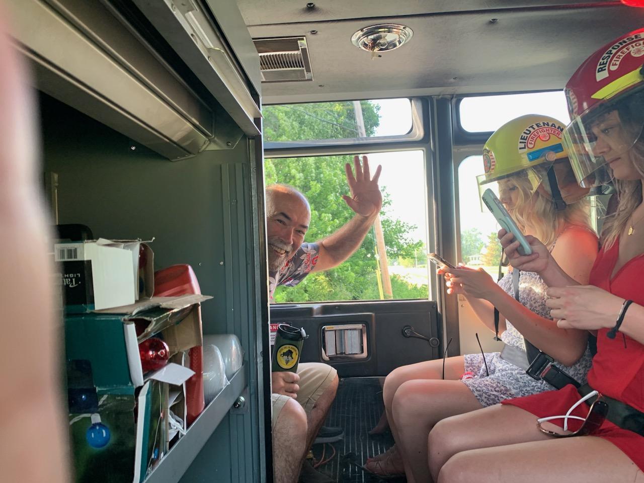 Person waving and smiling inside a bus with two women wearing firefighter helmets using phones.