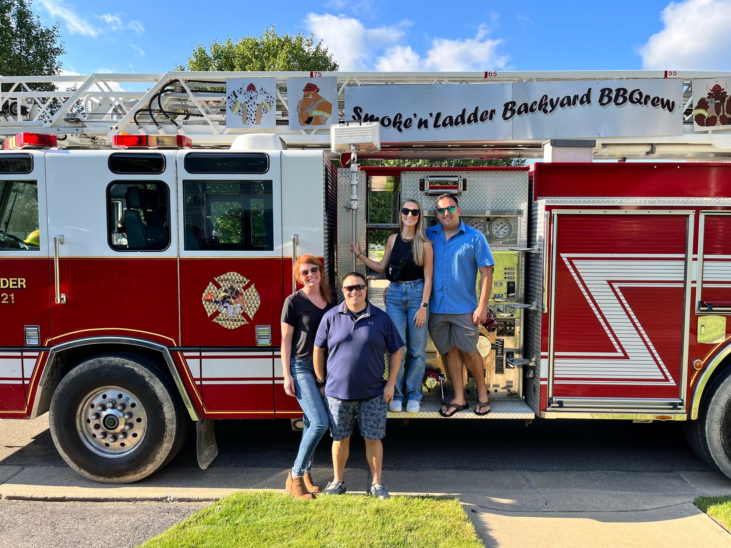 Three people standing in front of a red fire truck with a backyard BBQ theme, one person on the truck's step, outdoors.