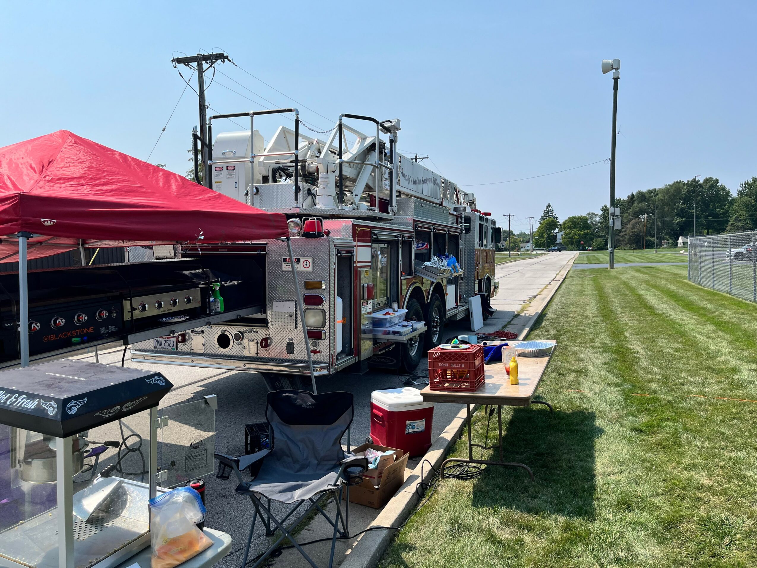 Fire truck parked on grass with a red canopy, table with supplies, and a portable grill, near a sports field.