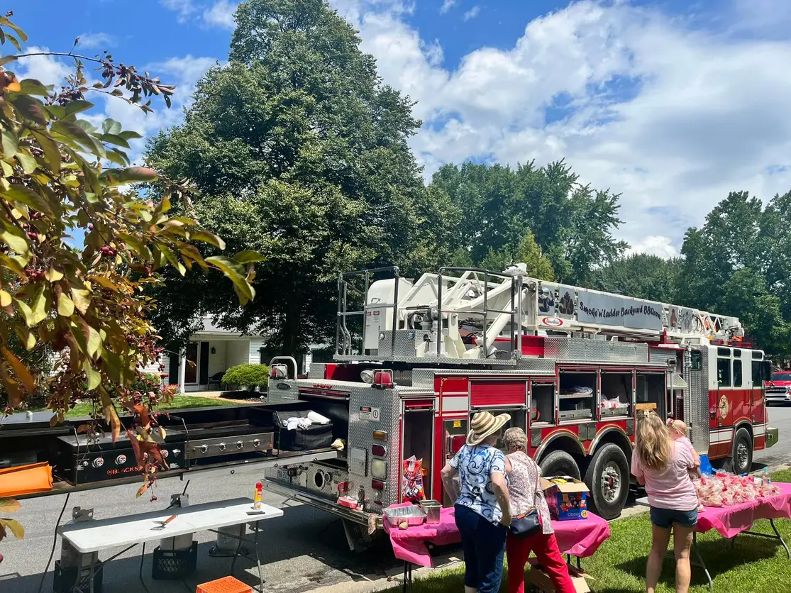 Fire truck parked outdoors with three people near pink tables, trees and blue sky in background.