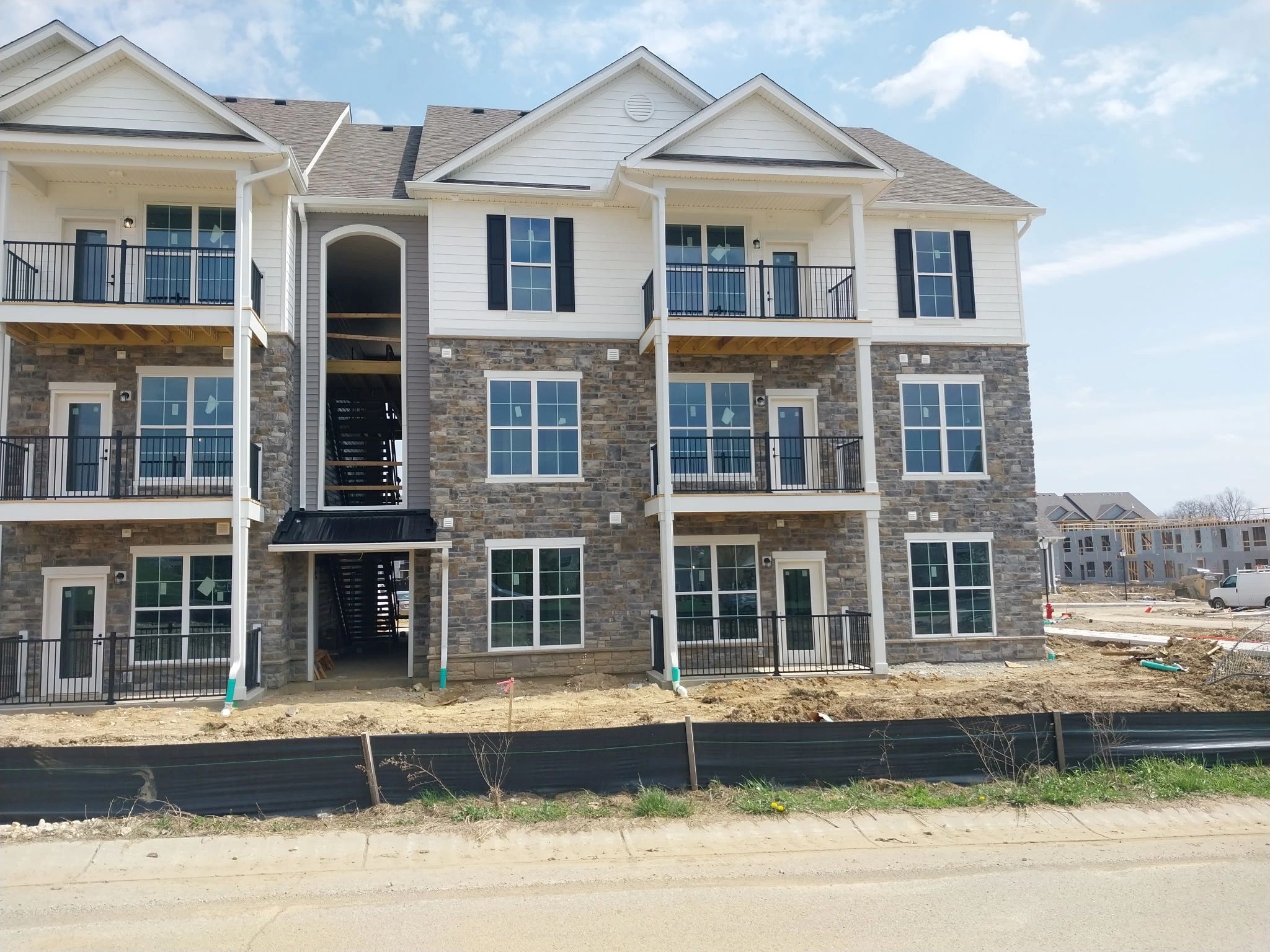 Three-story residential building with balconies, stone and siding exterior, under construction, with a dirt foreground and blue sky.