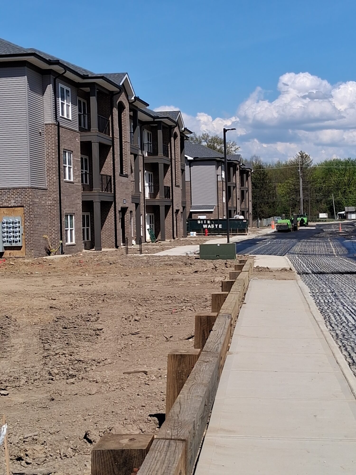 New apartment building with multiple floors and balconies, sidewalk, street, and parked cars under a blue sky.