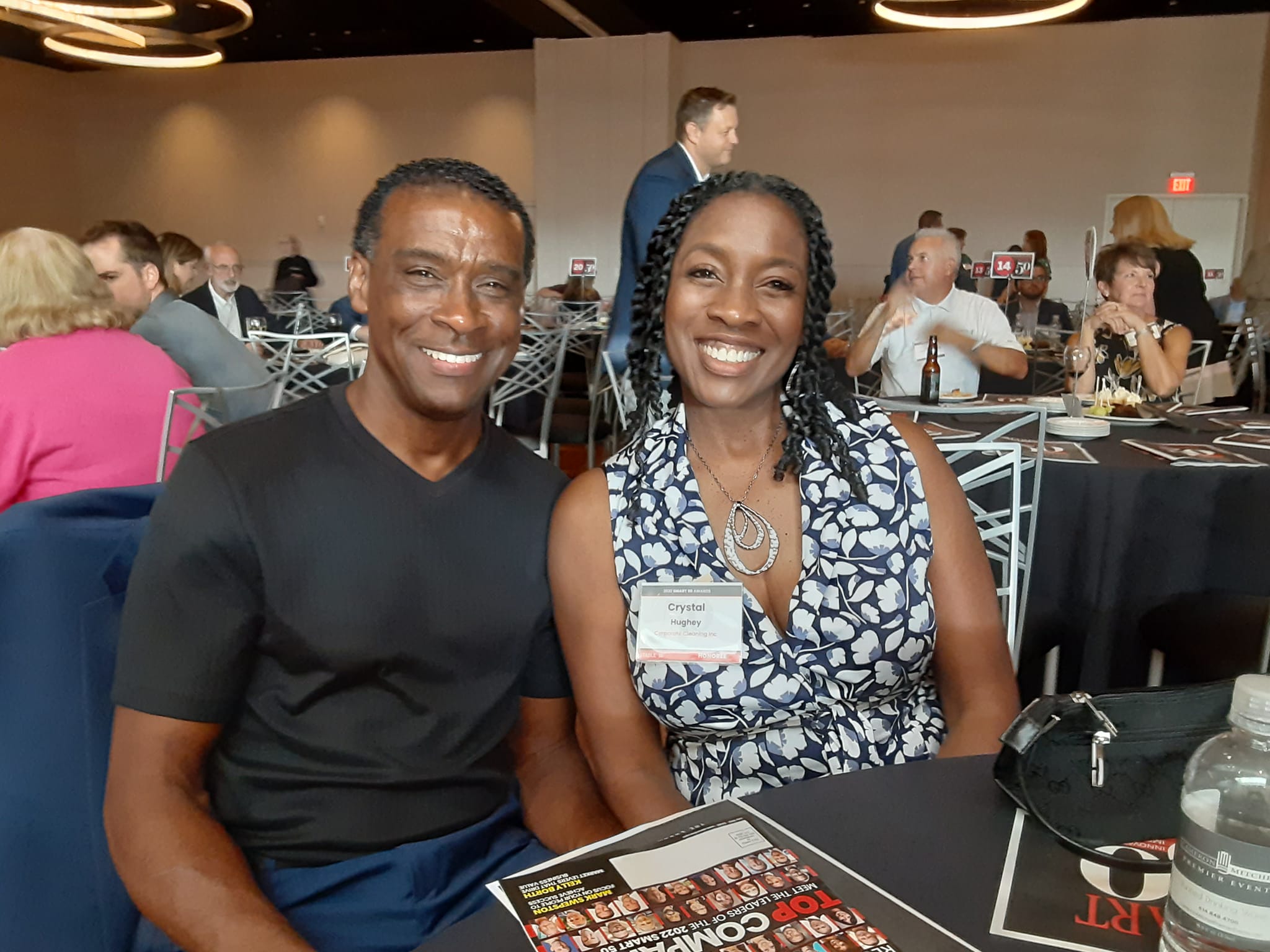 Two smiling people sitting at a table in a banquet hall, with other guests in the background.