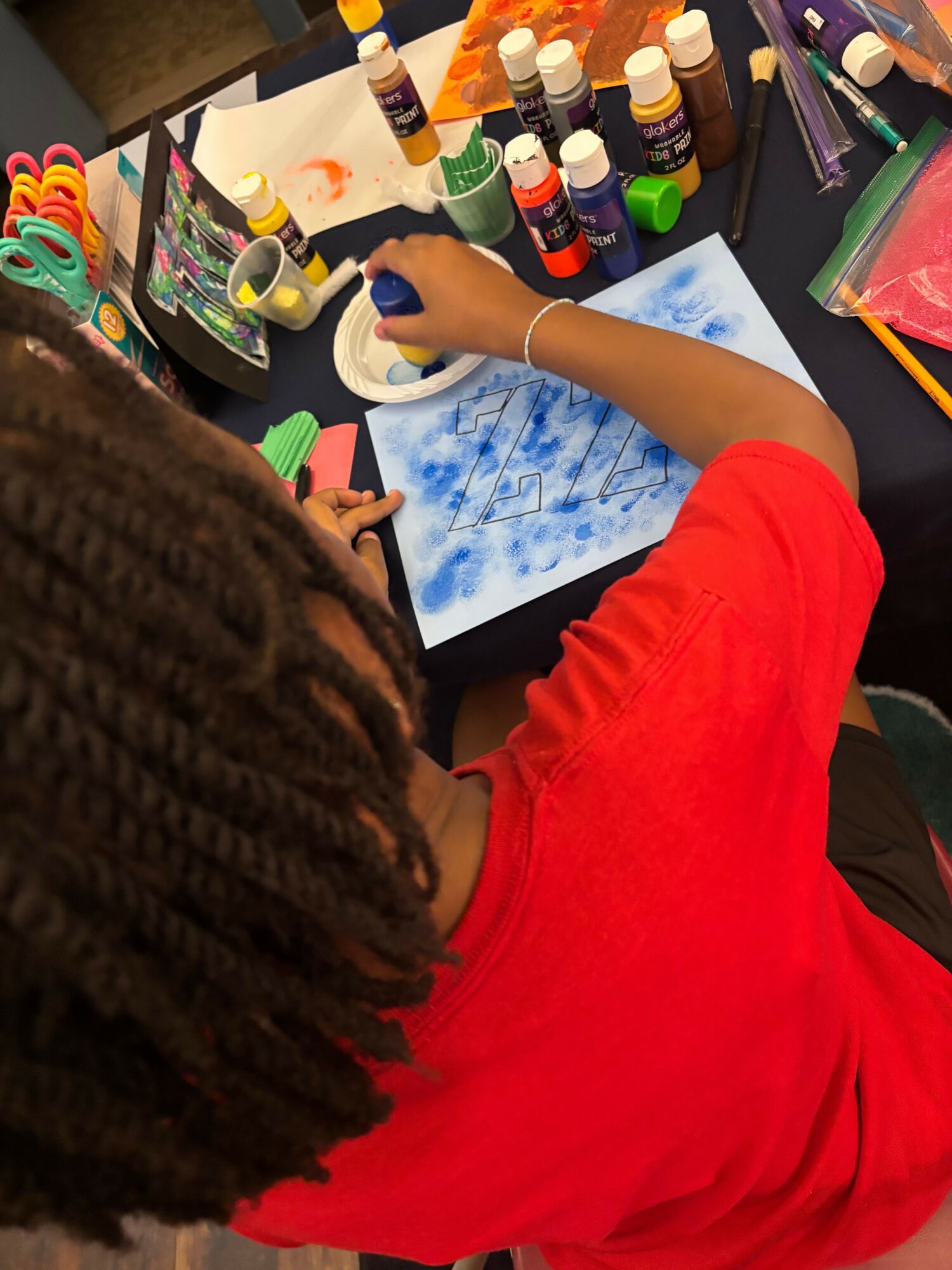 Child with braided hair in red shirt paints on paper with blue paint, surrounded by art supplies.
