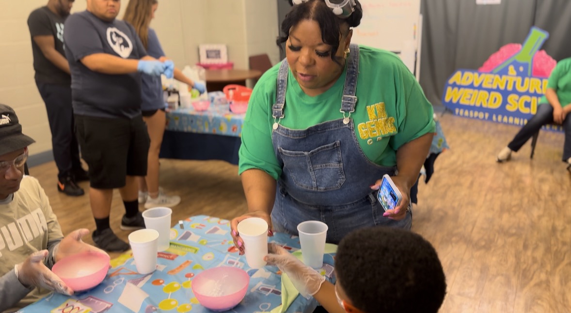 Woman in green shirt and apron pouring liquid into cups at a table with children.