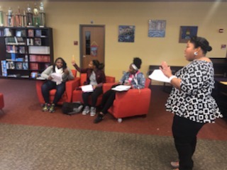 Four women sitting on red chairs and one woman standing in a room with bookshelves and paintings.
