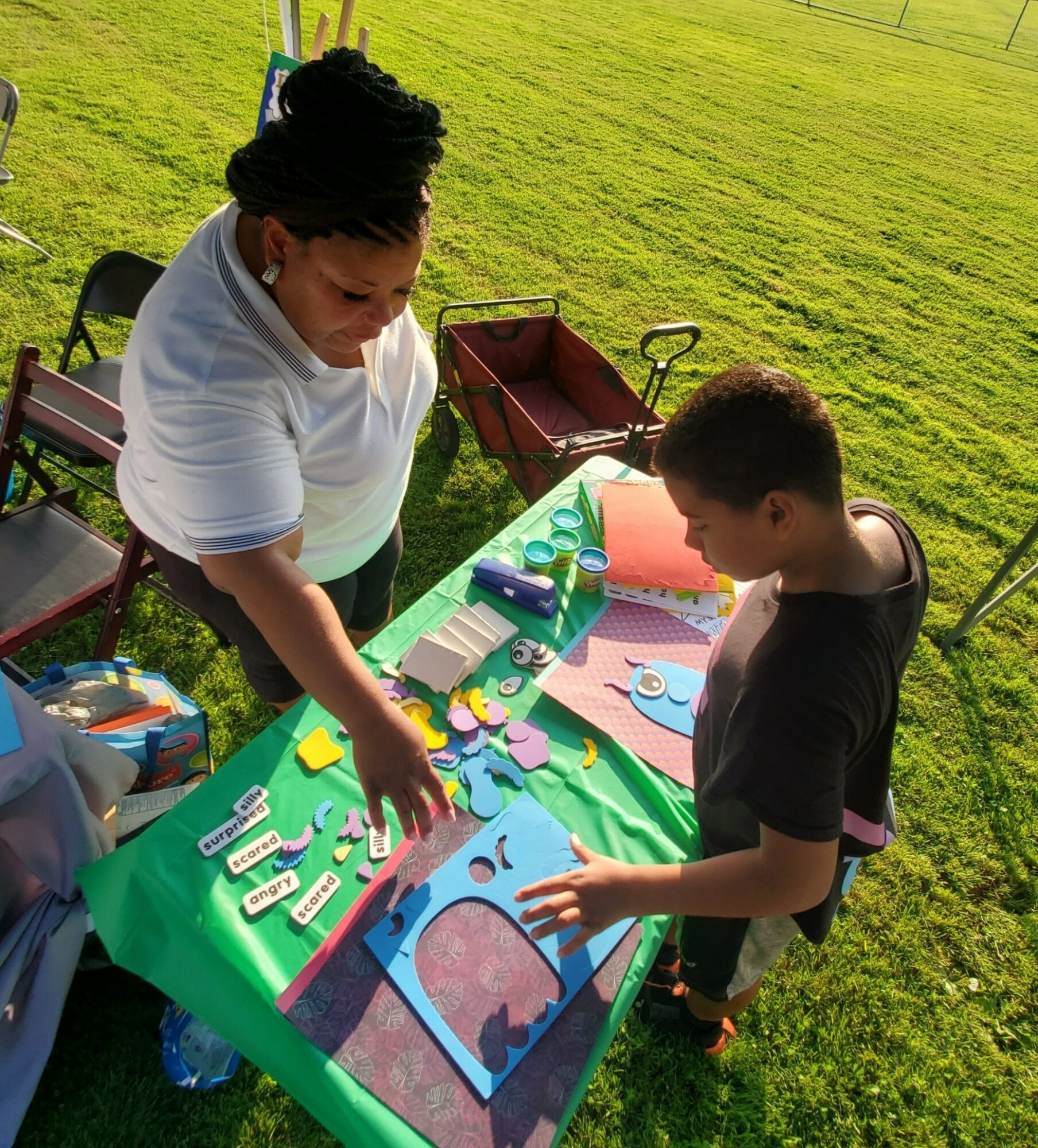 Two people working on a colorful art project on a green table outdoors, with grass and chairs nearby.
