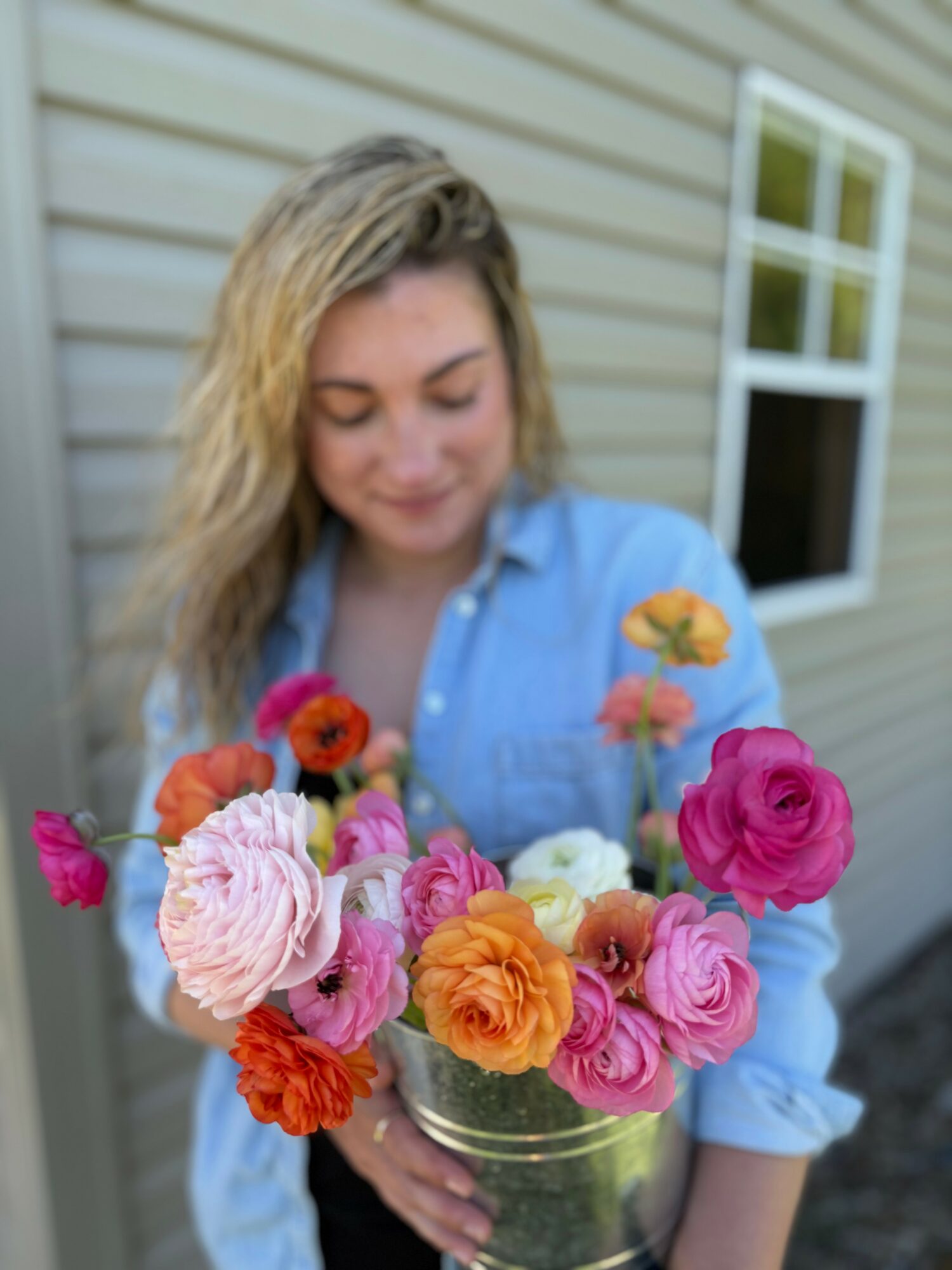 Woman holding a colorful bouquet of flowers outside a house with beige siding and a window.