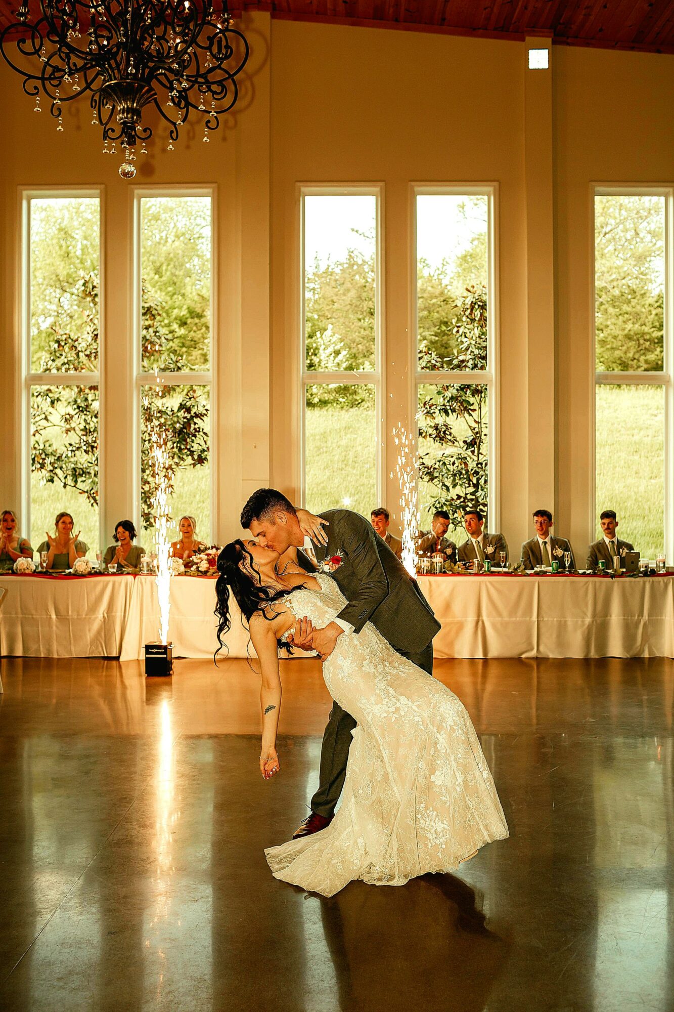 Couple dancing in a spacious room with tall windows and natural light, guests seated at tables in the background.