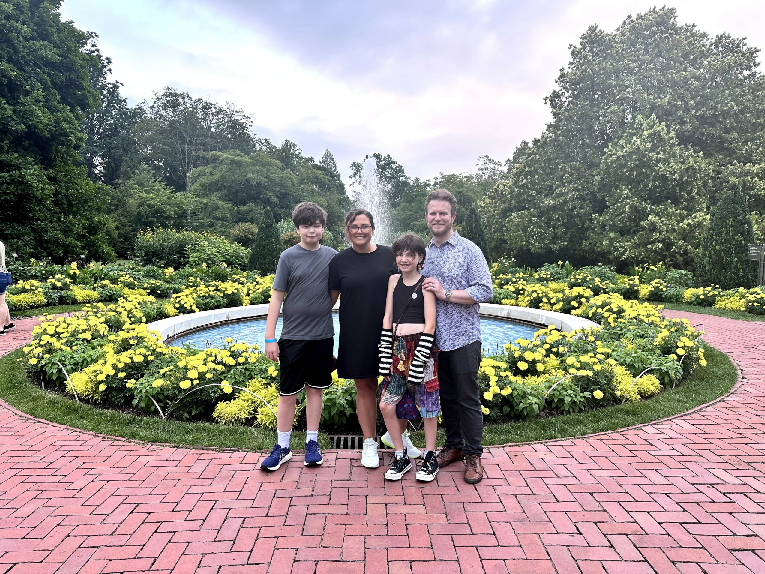 Four people standing in front of a circular flower bed with yellow flowers, trees, and a cloudy sky.