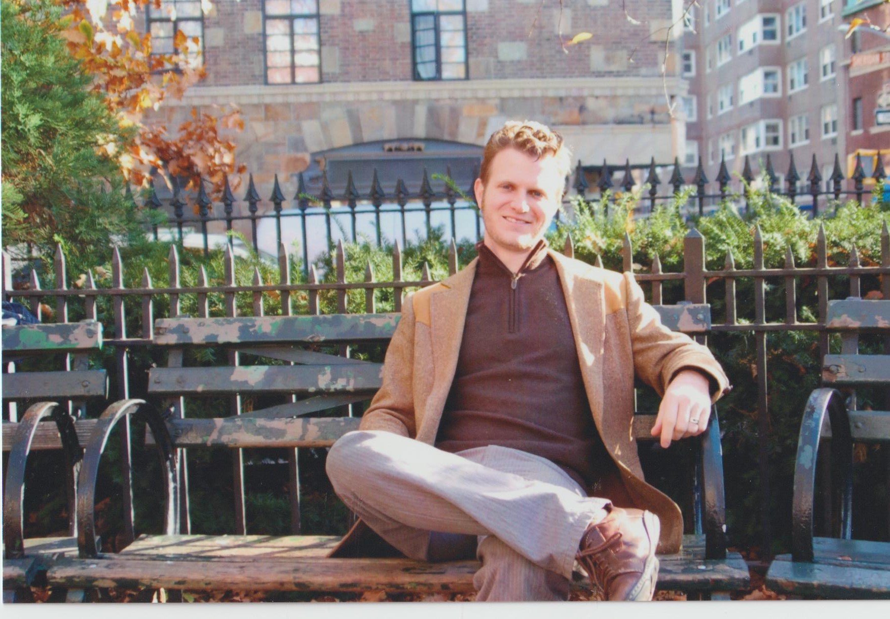 Young man sitting on a park bench with a backpack, smiling, in an urban park setting with buildings and trees.
