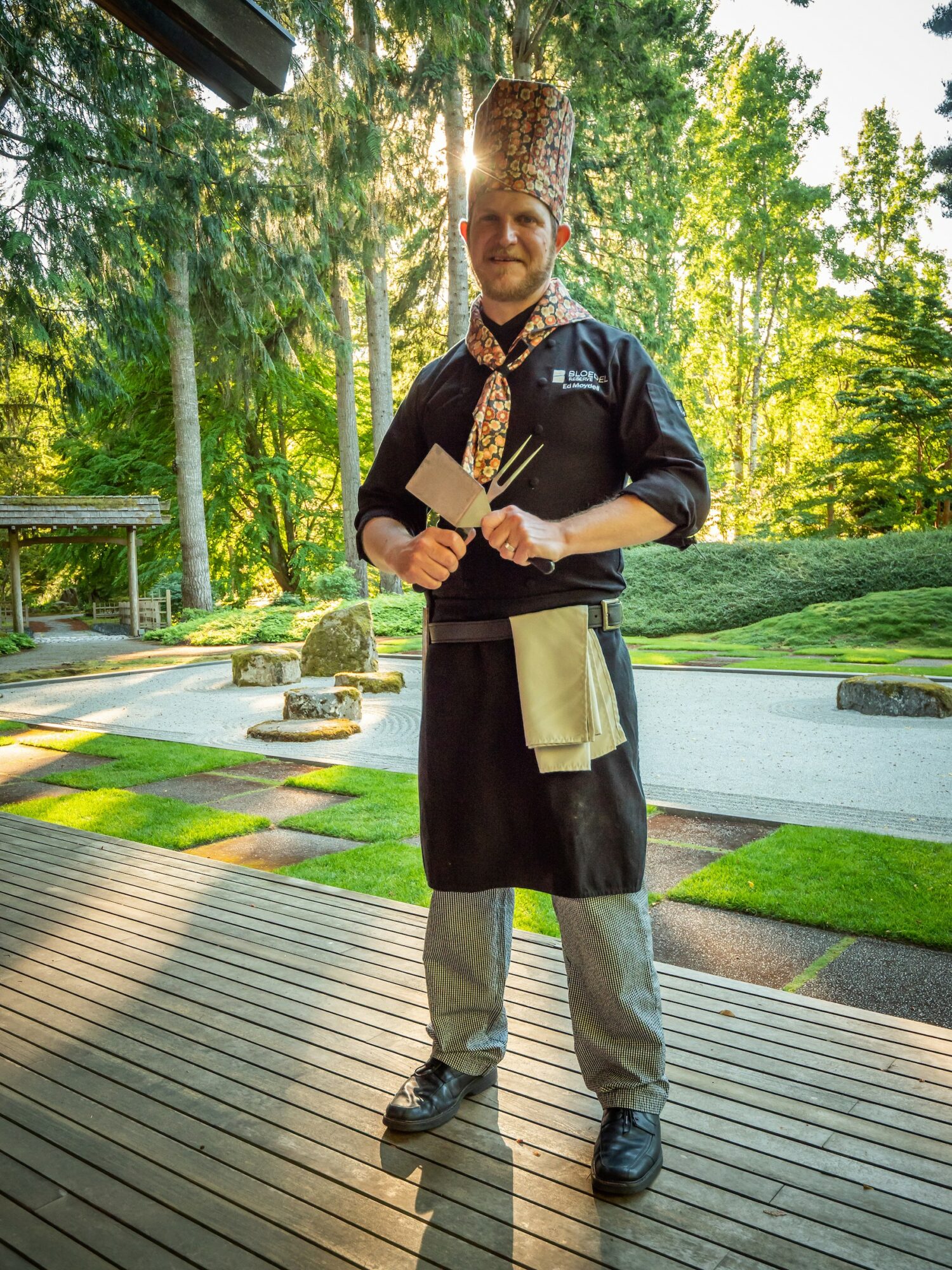 Person wearing a tall, patterned hat and traditional clothing, holding a wooden object, standing outdoors on a wooden deck.
