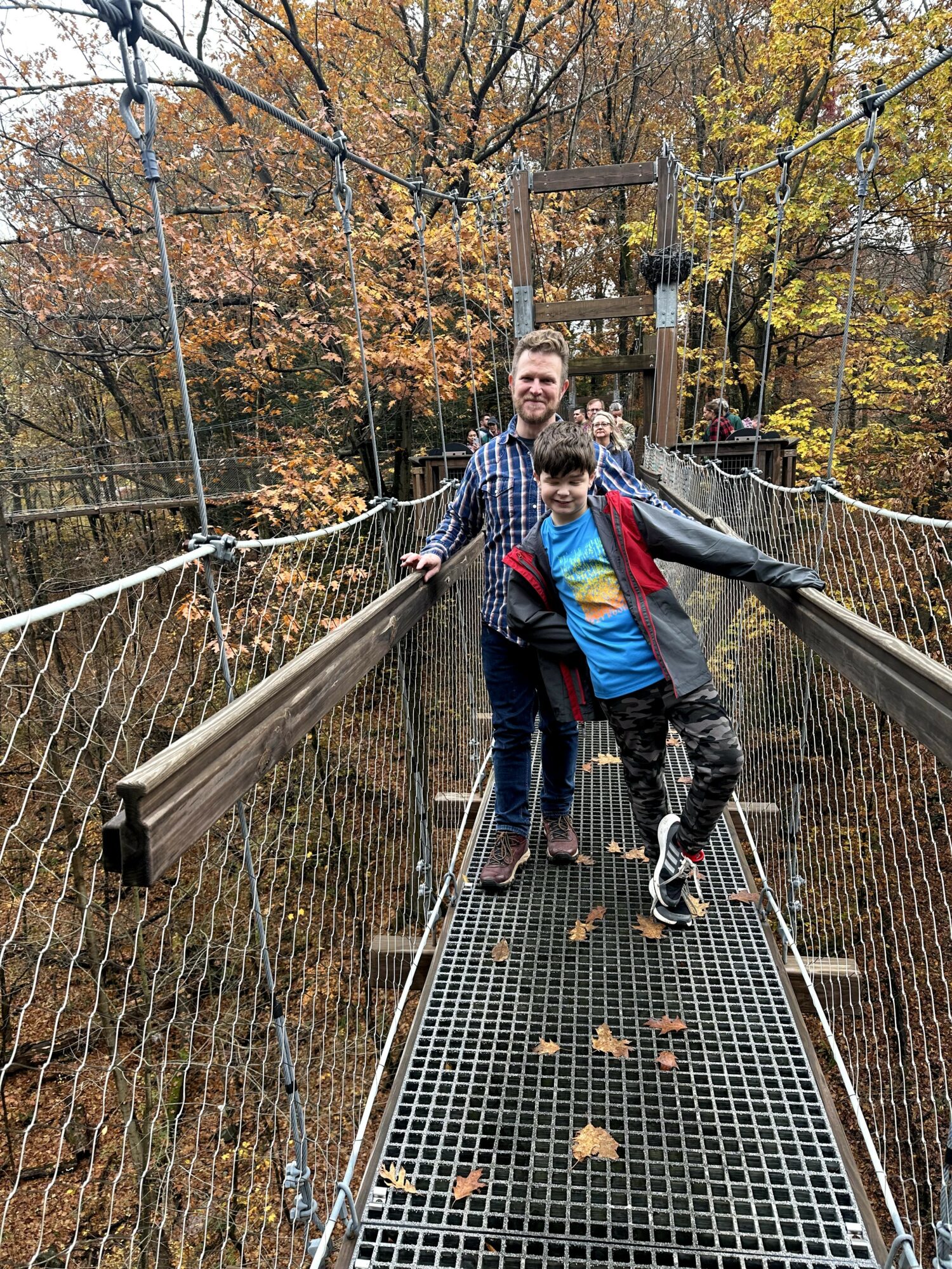 Two people walking on a suspension bridge in a forest with autumn leaves, one adult and one child, smiling.
