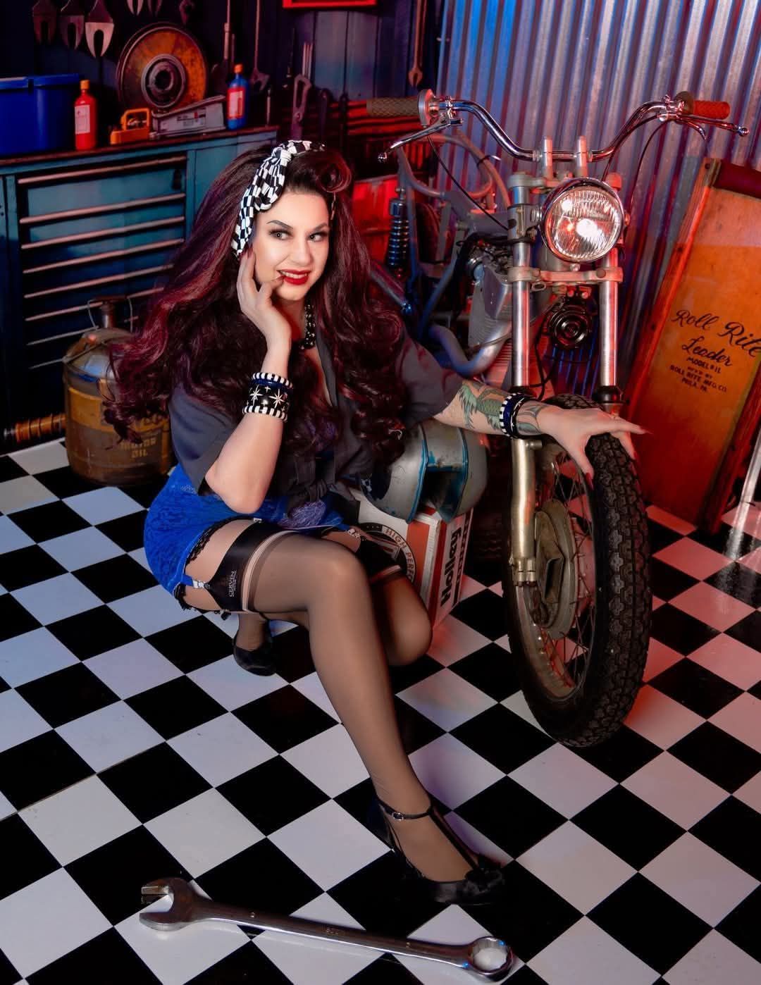 Woman with long dark hair, wearing a black top, denim shorts, and stockings, sitting on a motorcycle indoors.