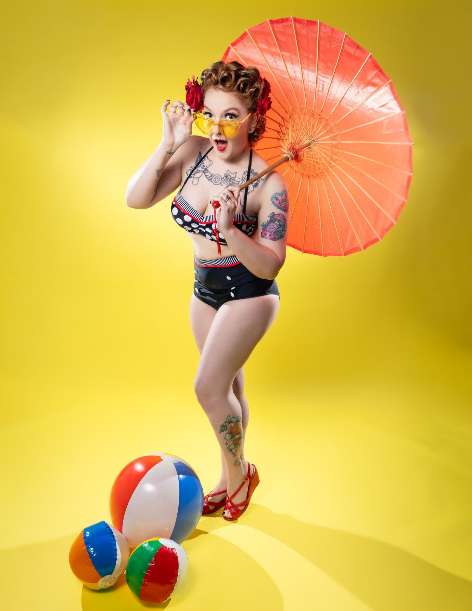 Person with tattoos, curly hair, sunglasses, holding an orange umbrella, standing on yellow background with beach balls.
