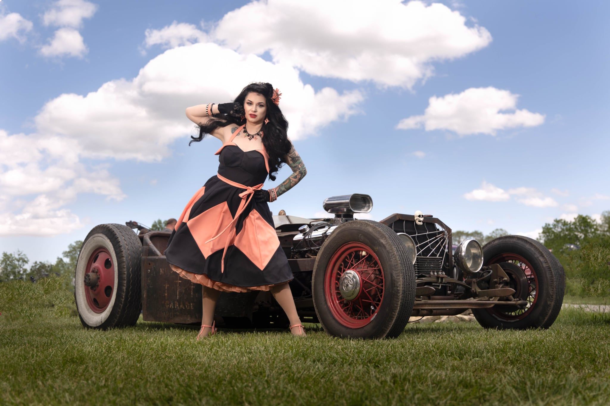 Woman in a dress standing next to a vintage race car outdoors with a grassy field and blue sky.
