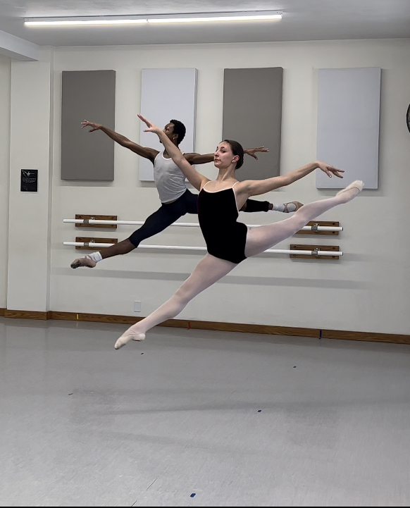 Two ballet dancers perform jumps in a dance studio with white walls and acoustic panels.
