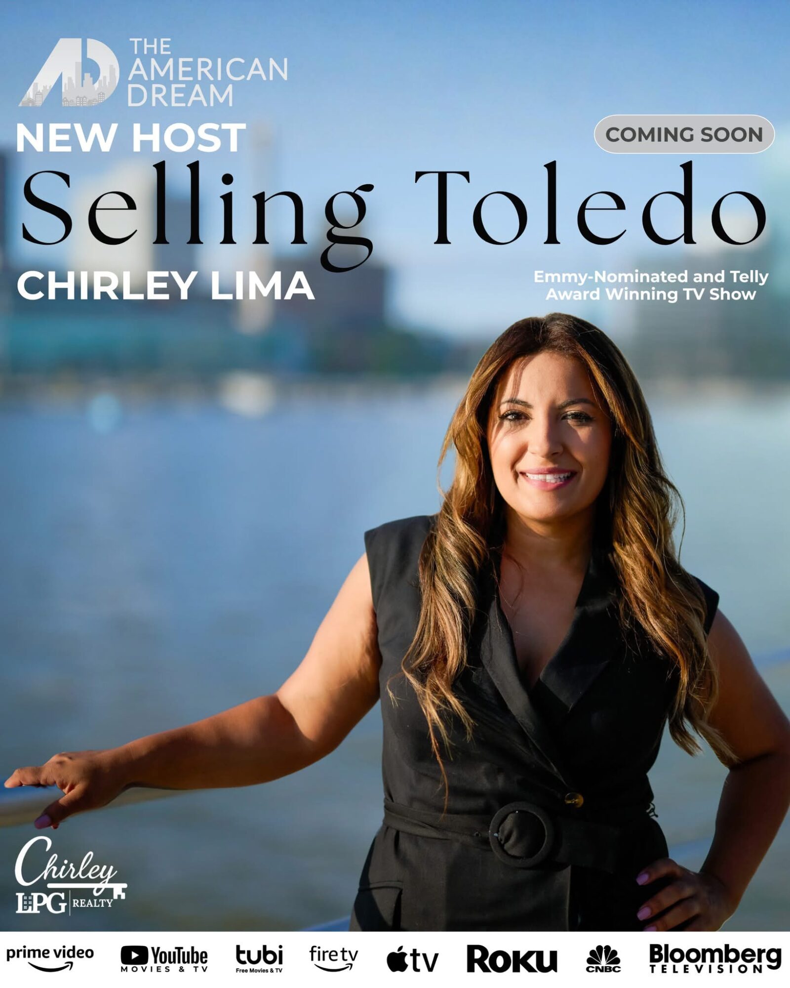 Woman with long hair smiling outdoors near water, wearing a black sleeveless top, with cityscape in background.