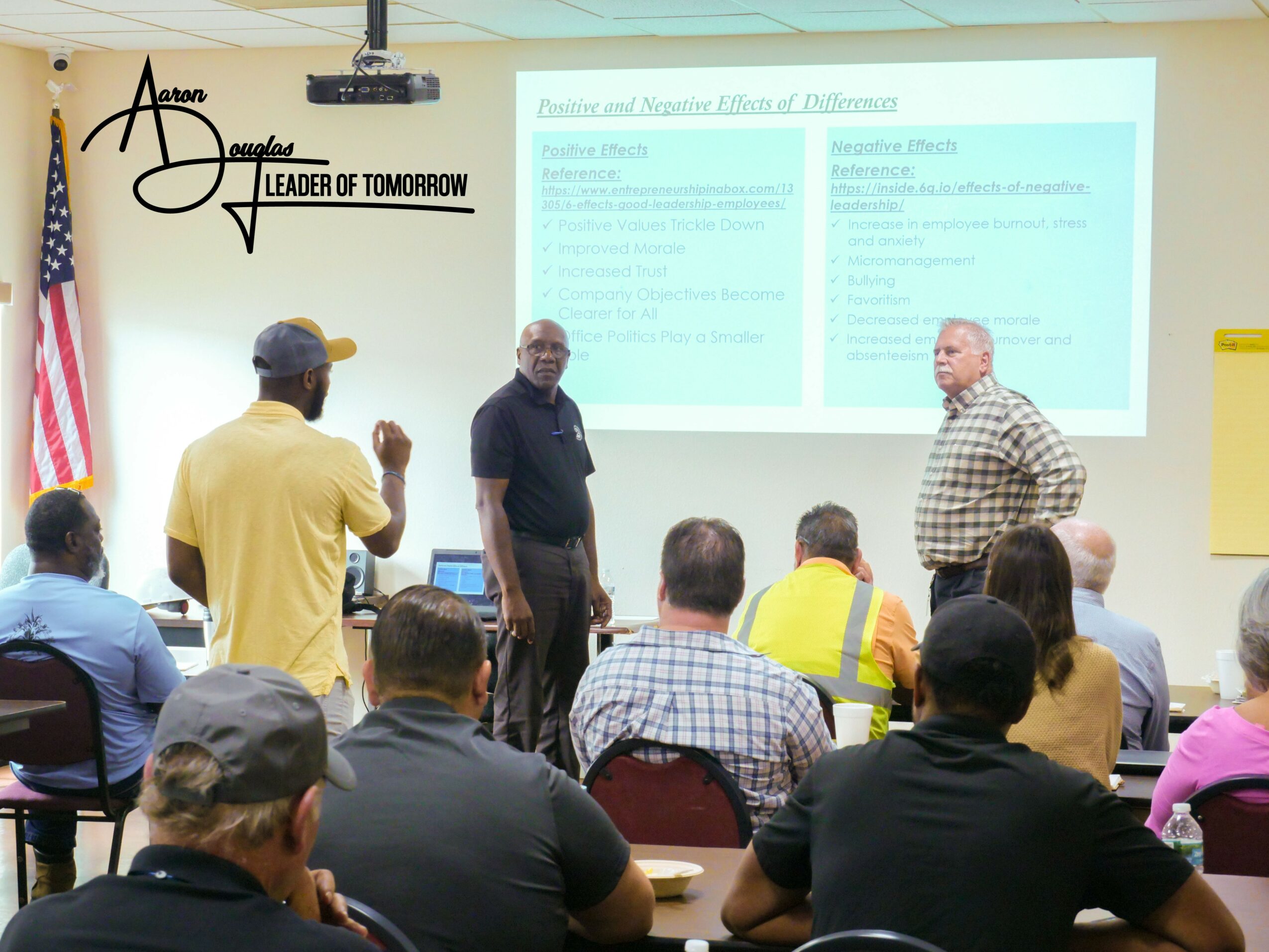 Classroom with people listening to two speakers at the front, presentation slide on the wall, American flag nearby.