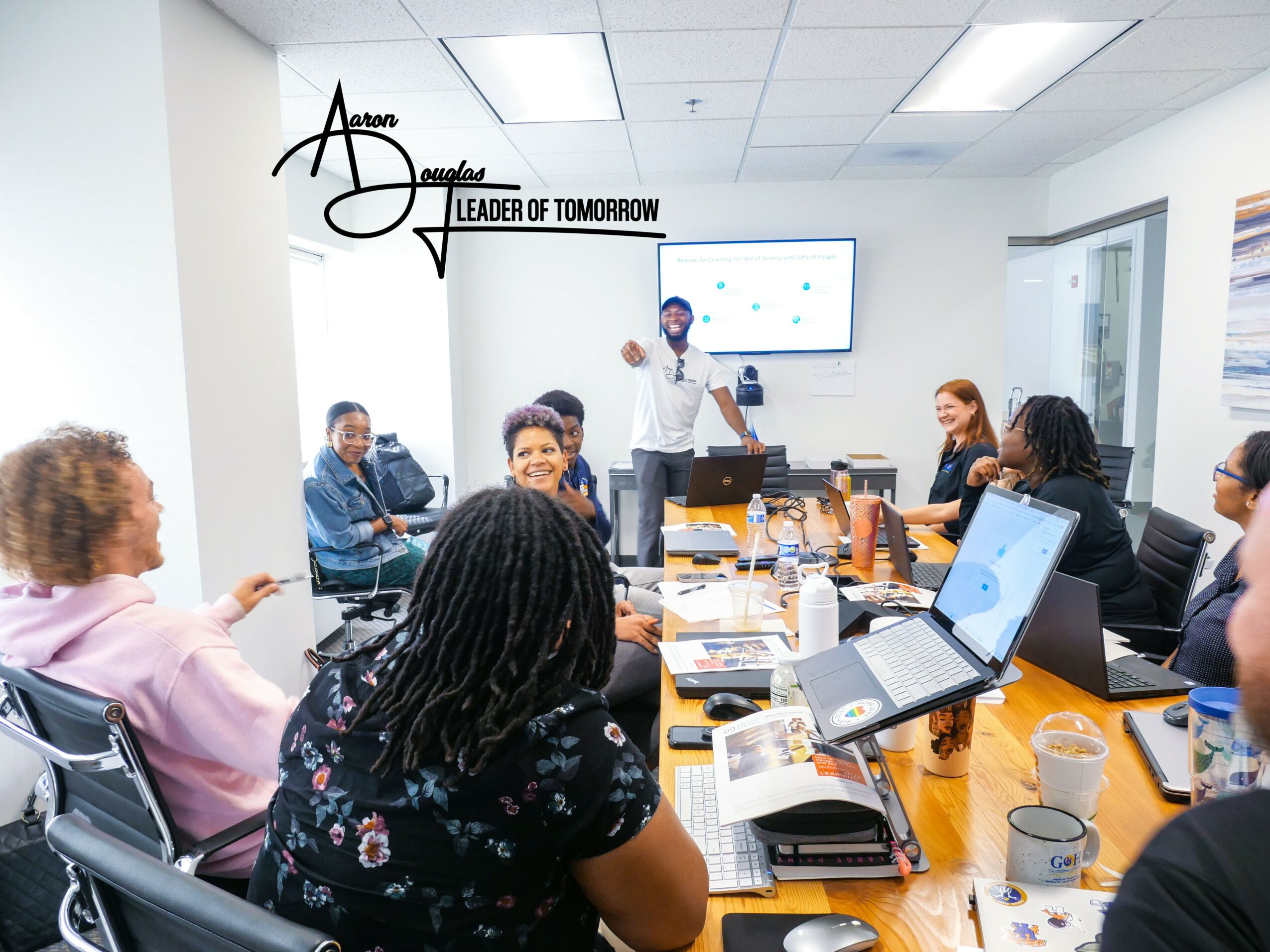 Meeting room with people seated around a table, a presenter standing, screen on wall, laptops and papers on table.