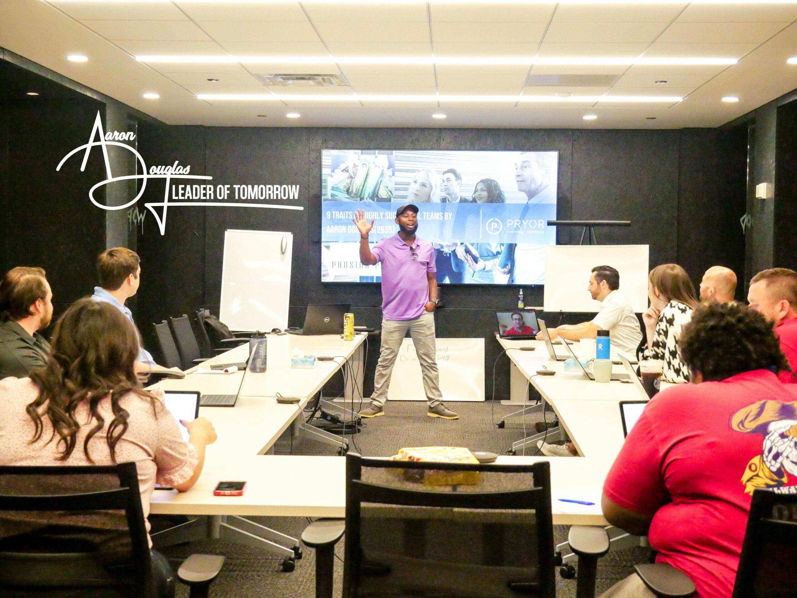 Person standing in front of a screen, speaking to a group in a conference room with tables and laptops.