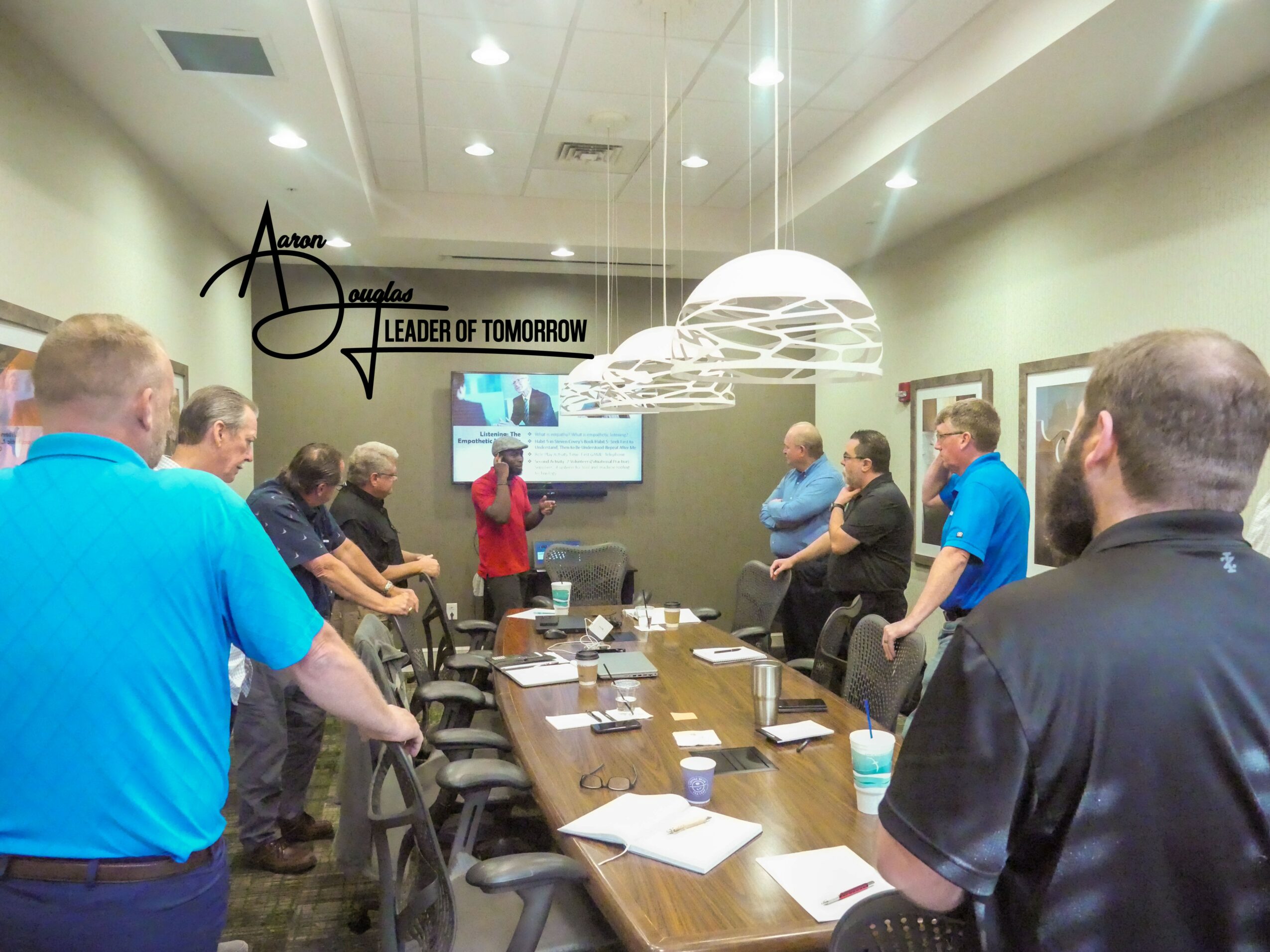 Group of people in a conference room listening to a presenter near a screen, with a long table and papers.