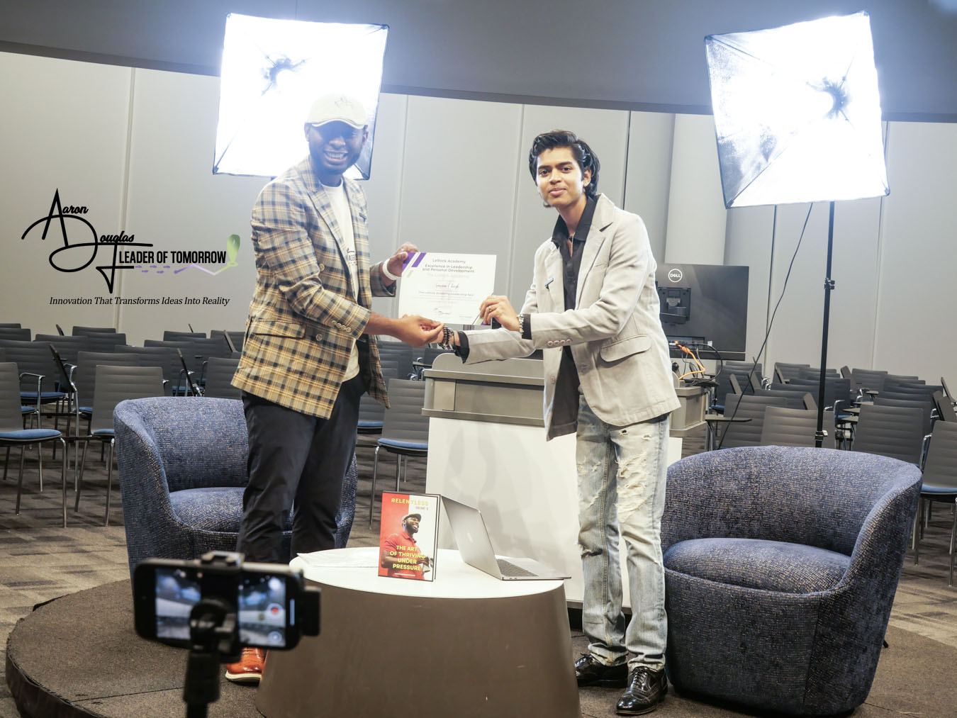 Two men exchanging a certificate in a conference room with chairs, two large lights, and a table with a book.