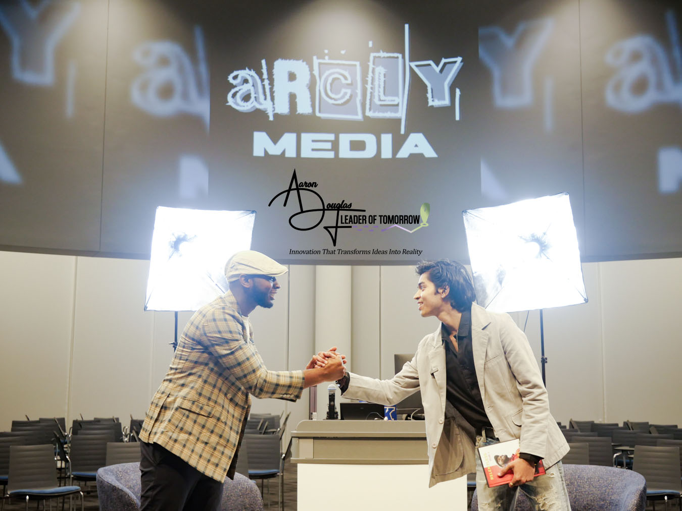 Two people shaking hands on stage with bright lights and a large screen behind them displaying logos and text.