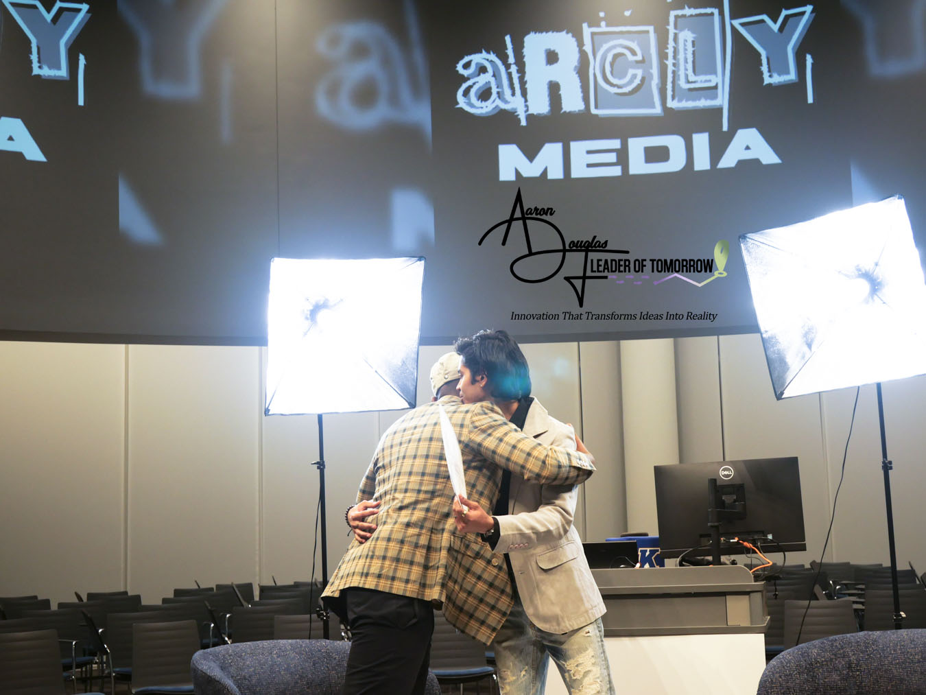 Two people hugging in a conference room with media equipment and bright lights, logo on screen behind them.