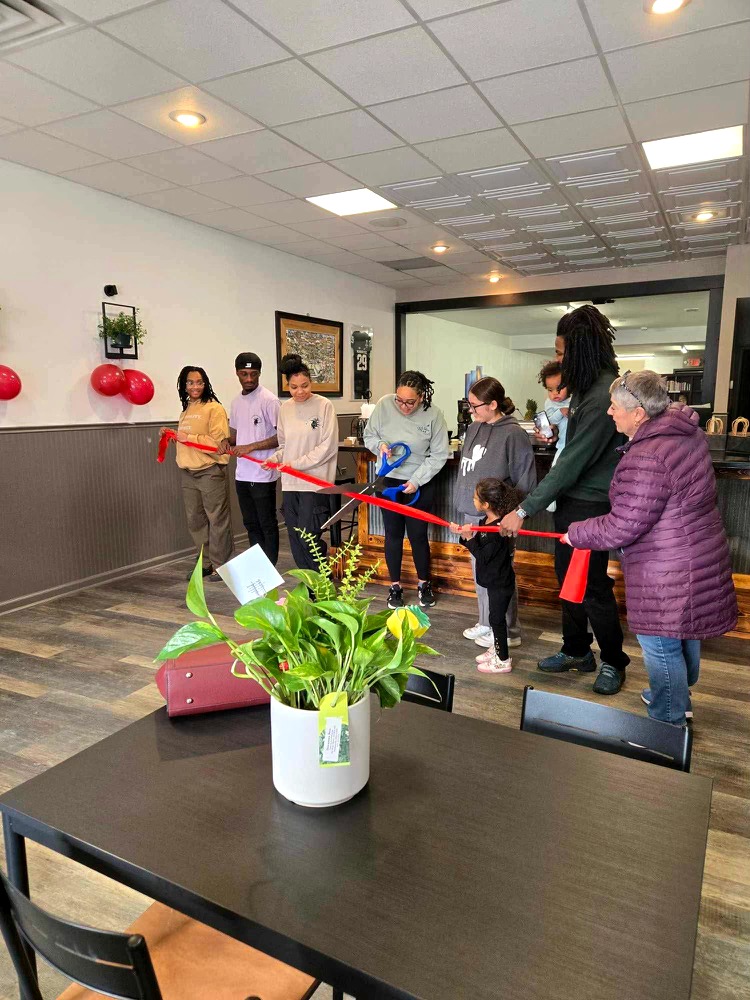 Group of people participating in a ribbon-cutting ceremony indoors, with a potted plant on a table in foreground.