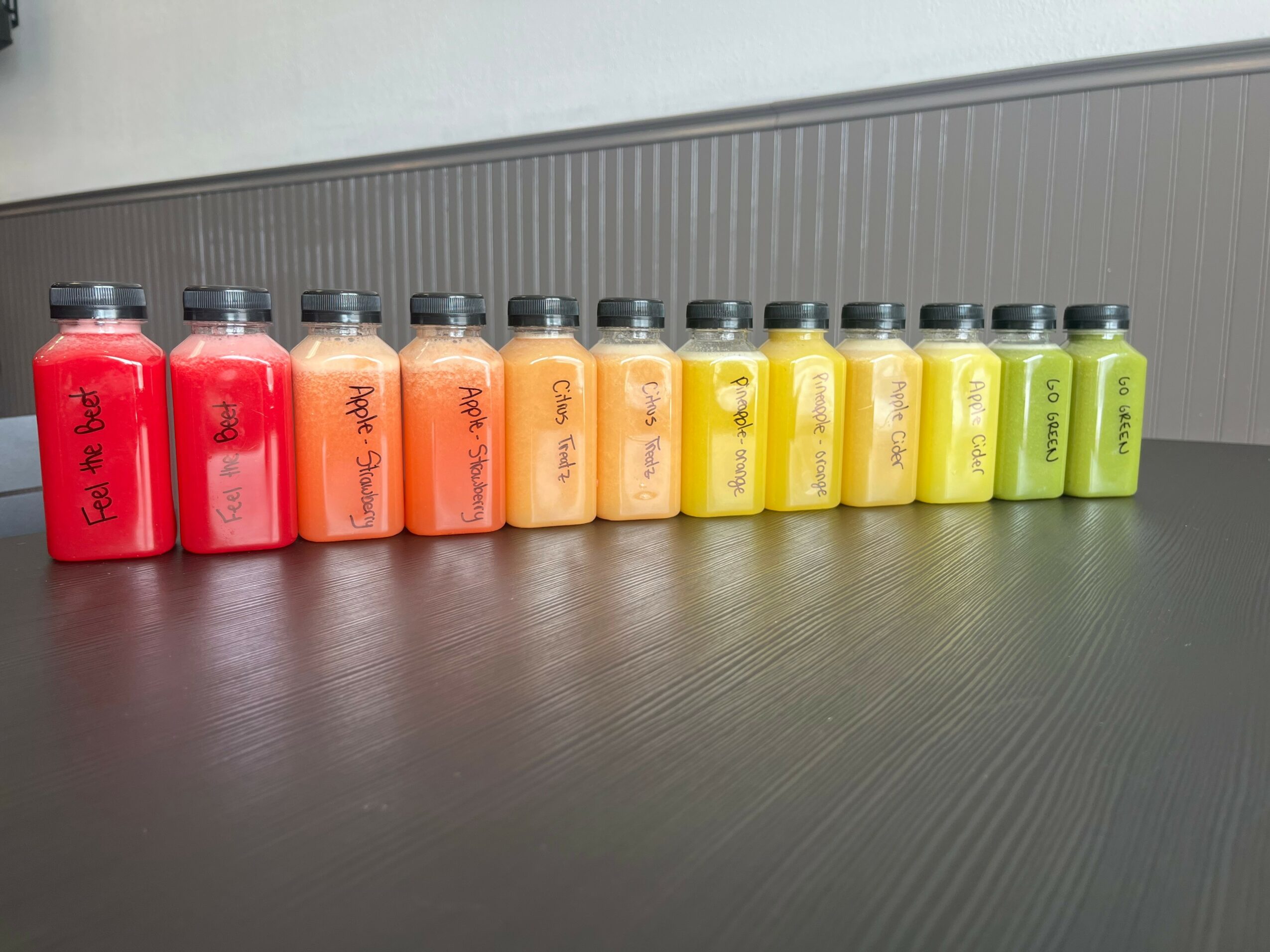 Row of ten small bottles with black caps, containing colorful liquids from red to green, on a black surface.