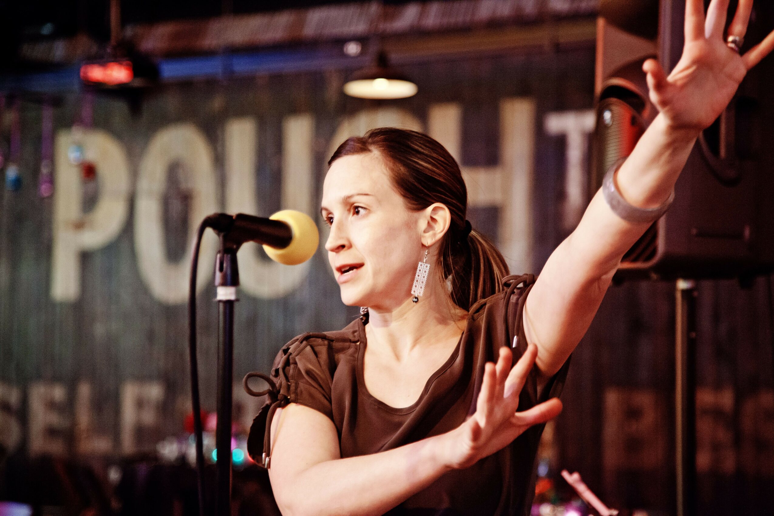Woman speaking into microphone with one arm raised, in a dimly lit venue with a sign in the background.