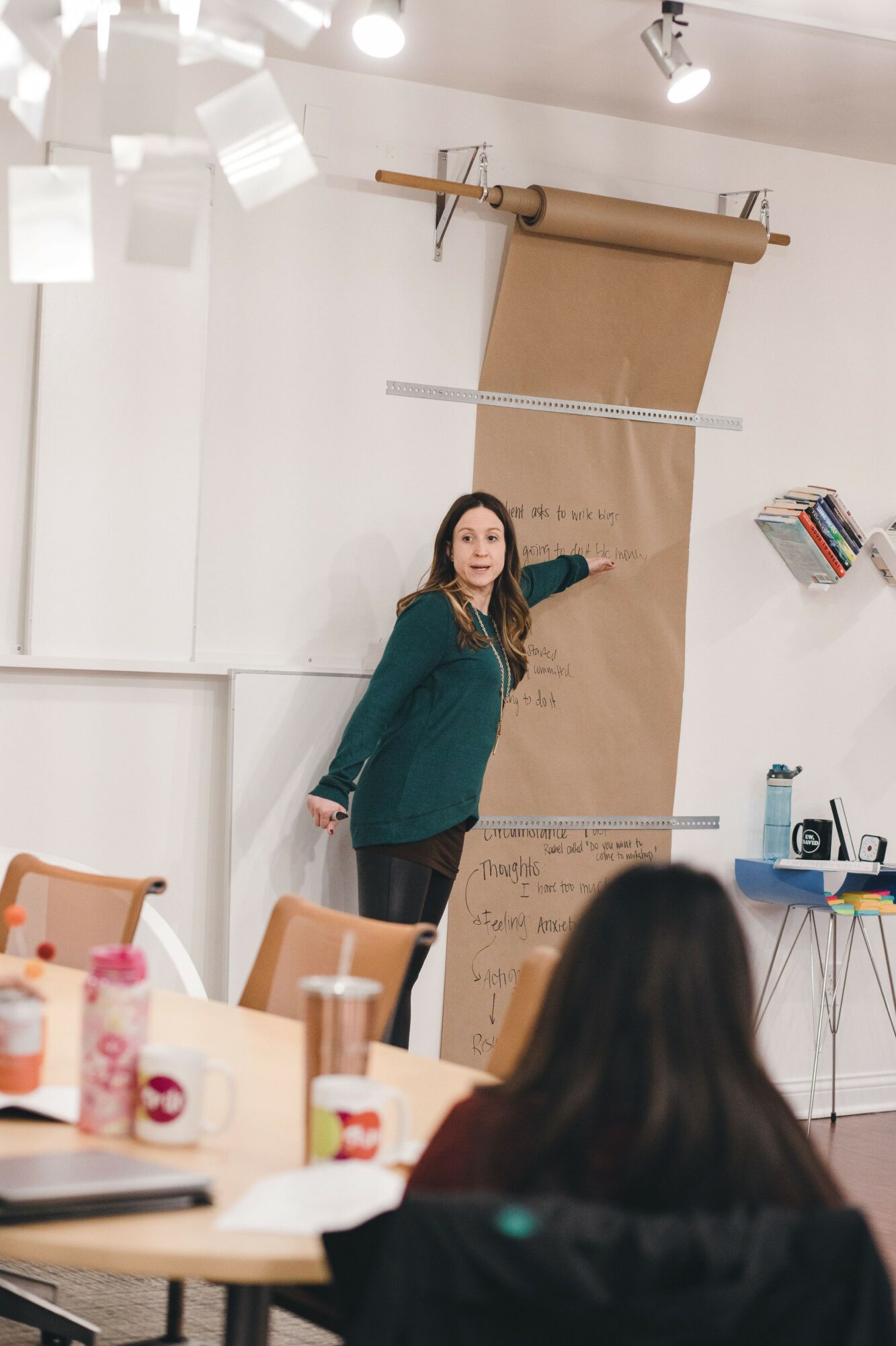 Woman in green jacket standing next to a large paper scroll on wall, pointing at it in a room with chairs and tables.